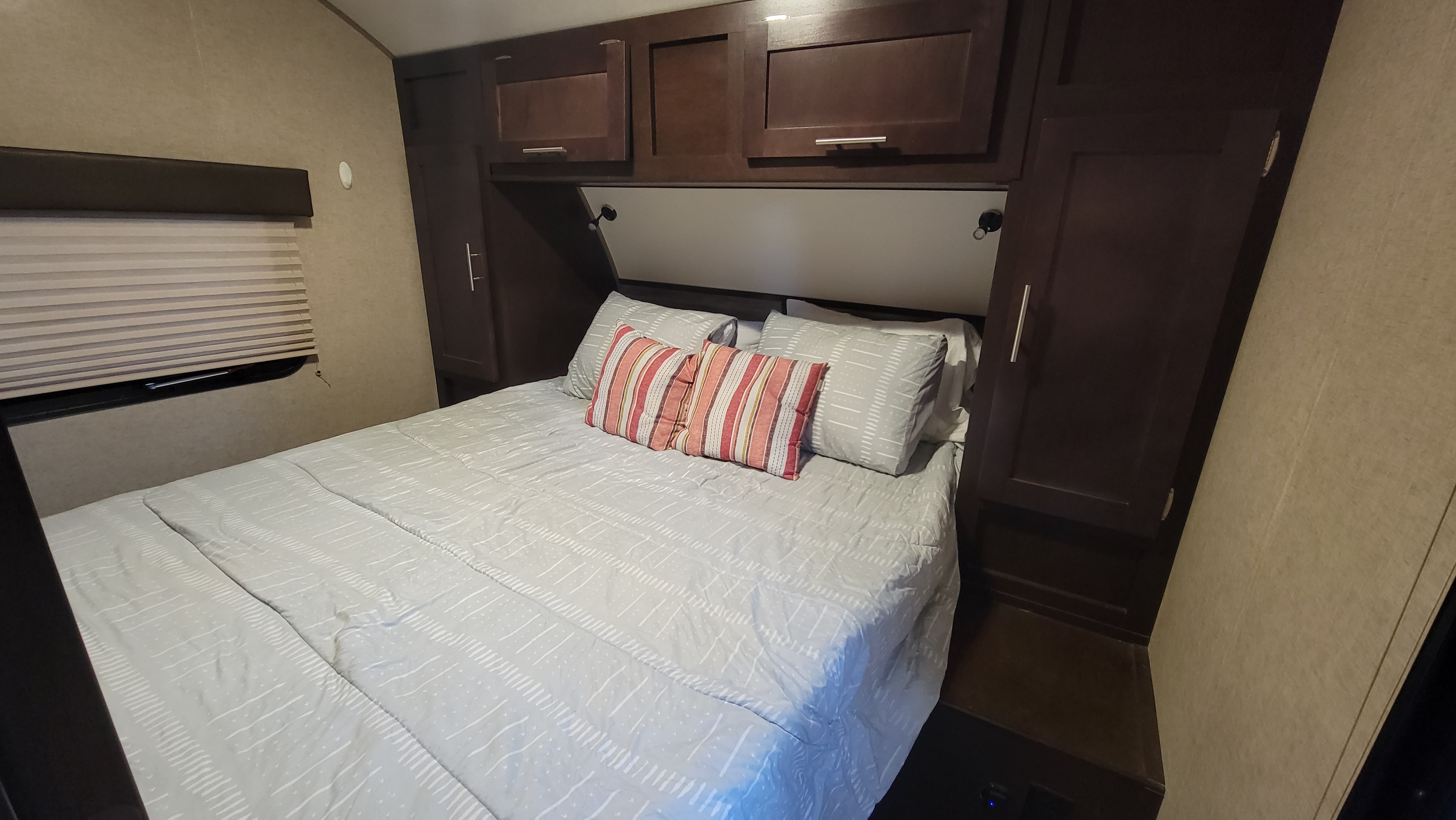 Cozy RV rental bedroom featuring a bed with striped pillows, dark wood overhead cabinets, and a window with blinds.. Forest River Cherokee 2019
