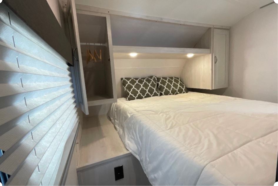 Inside an RV rental: a compact bedroom with bed, two patterned pillows, overhead cabinets, and window blinds.. Forest River Prime Time Tracer 2021