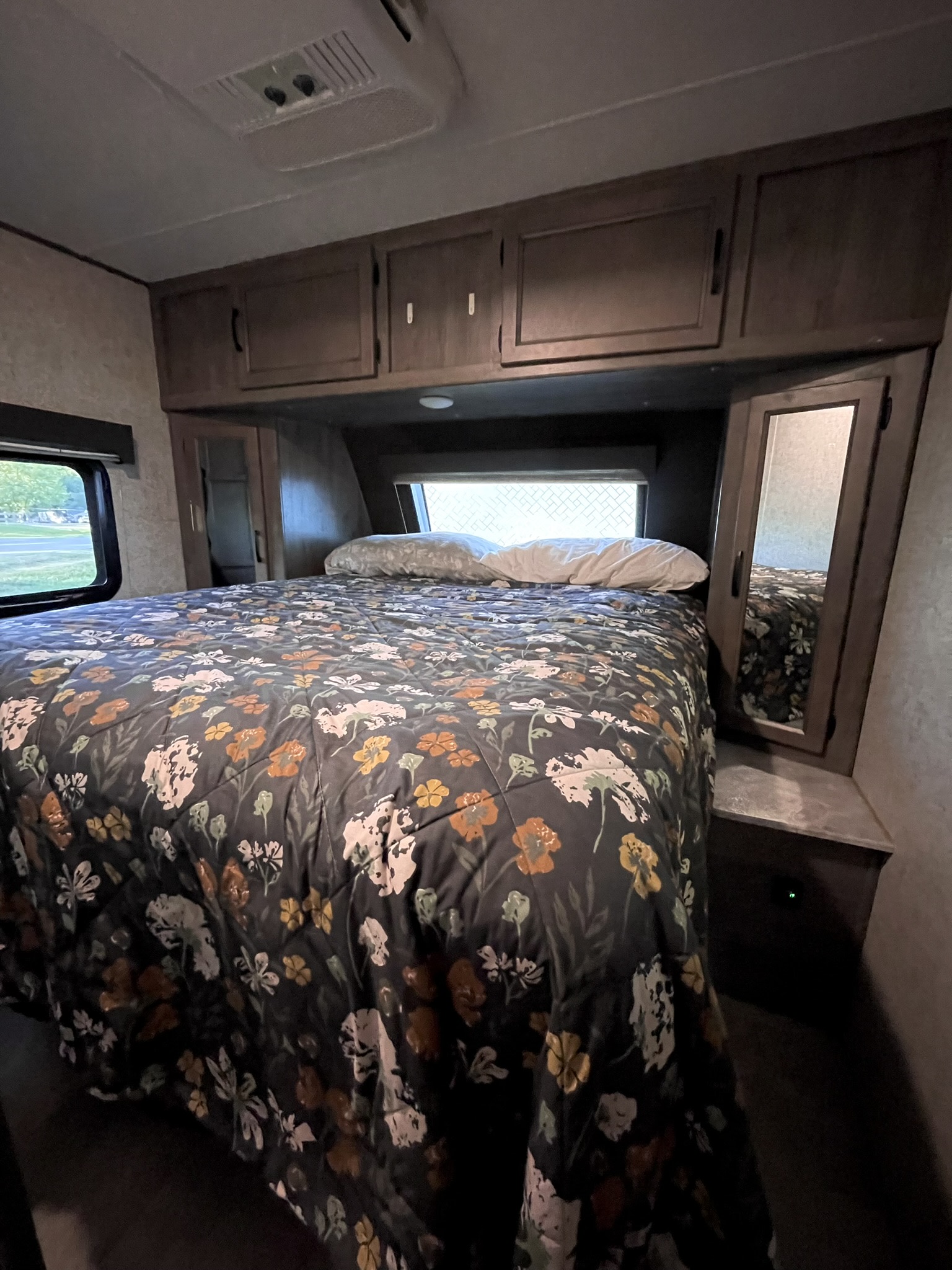 Inside an RV rental, a comfy bedroom features a floral bedspread, ample storage cabinets, and a side window.. Coachmen Apex 2020