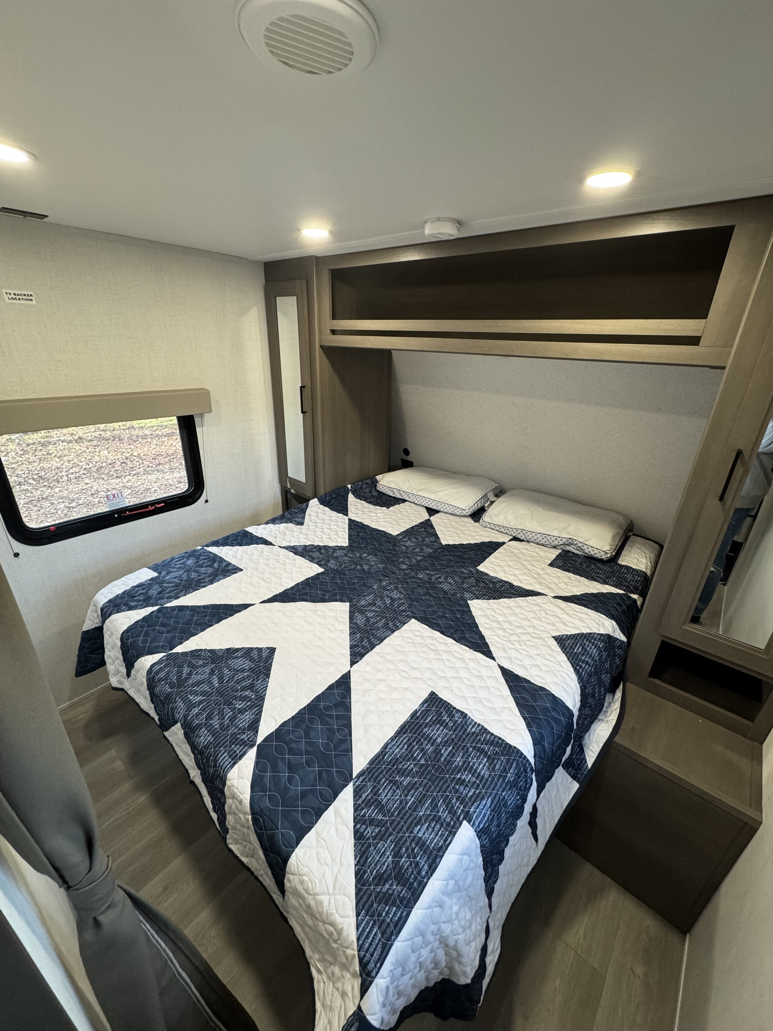 Cozy RV rental bedroom with a star-patterned quilt, overhead storage, and window. Perfect for your next trip.. Grand Design Transcend 2025