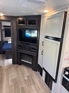 RV rental living space: dark entertainment unit with TV & fireplace, beside a tall refrigerator. Bedroom area visible.. Jayco White Hawk 2022