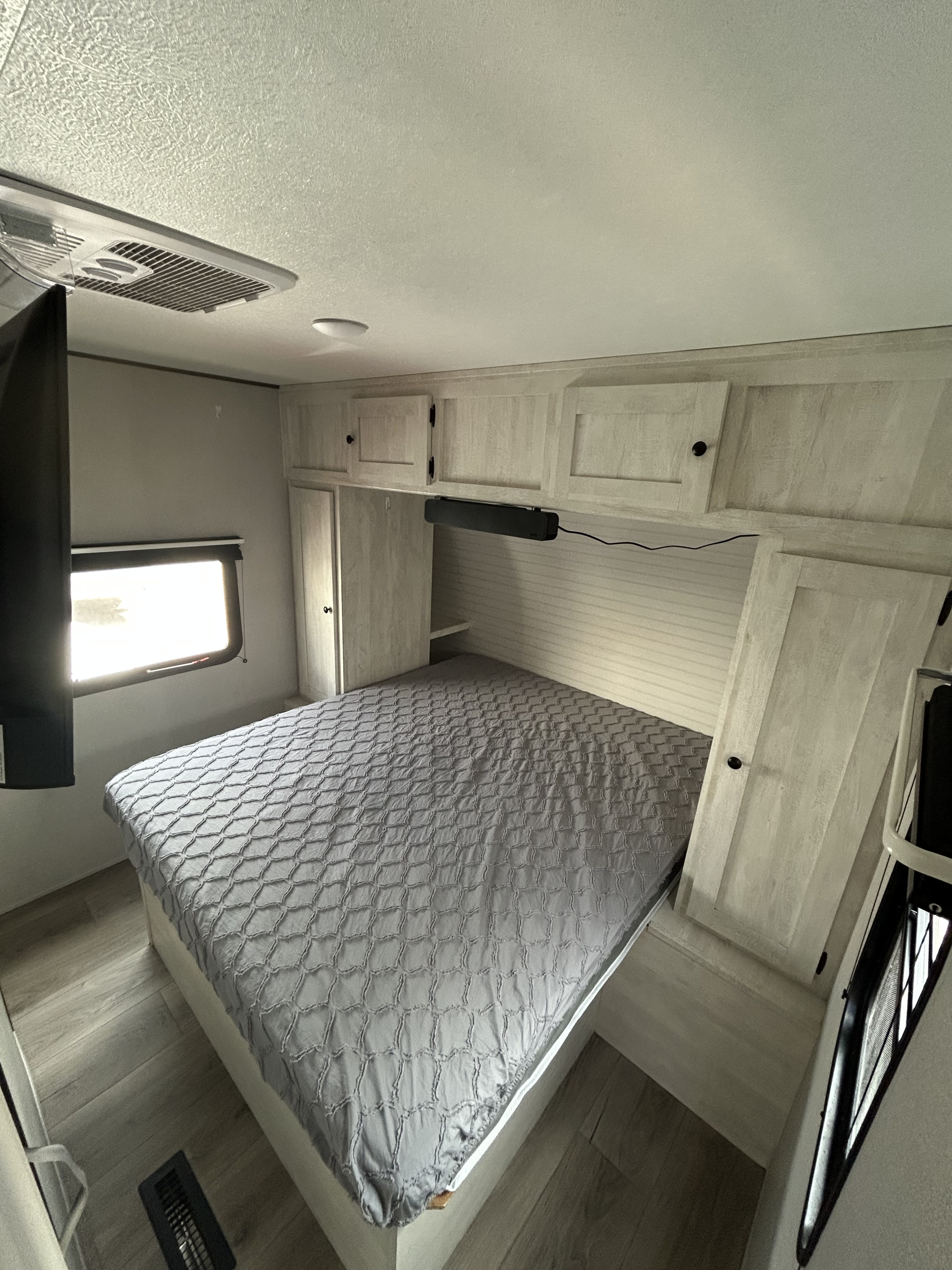 Bright RV rental bedroom features a comfy bed with a grey textured comforter, light wood-look cabinets, TV, & window.. Forest River Shasta i-5 2024
