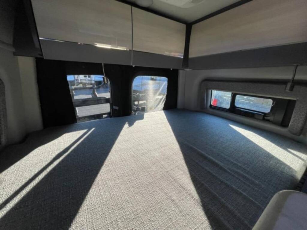 Inside a modern RV rental: a large bed fills the floor, surrounded by windows and overhead storage cabinets. Natural light streams in.. Mercedes Sprinter 2022