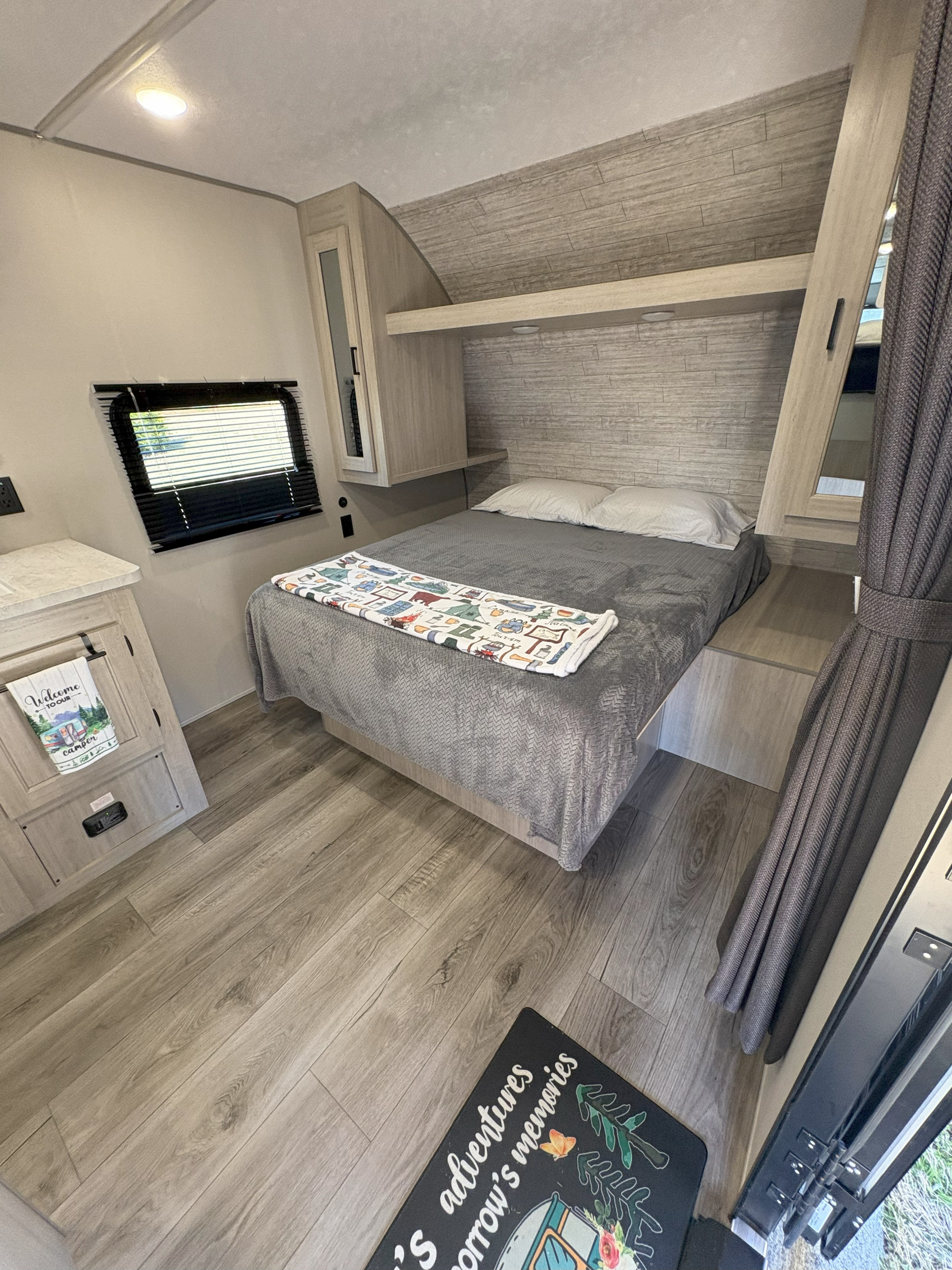Step into this cozy RV rental bedroom, featuring a bed, built-in storage, wood-look flooring, and a window with blinds.. Coachmen Coachmen Catalina Summit 2025