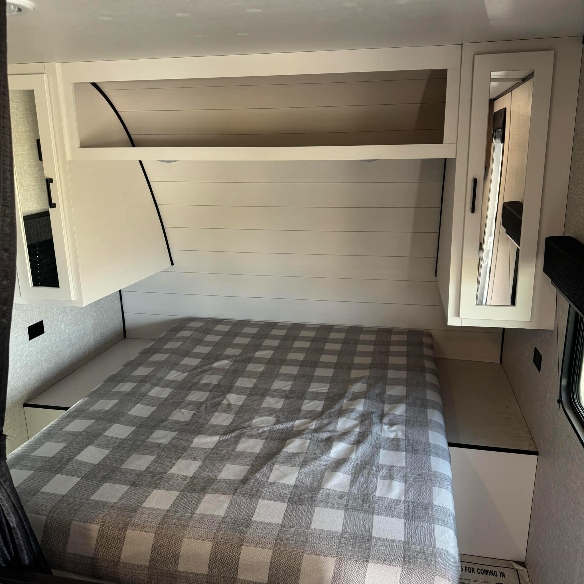 Interior of an RV rental bedroom featuring a gray plaid bed, white shiplap wall, and overhead storage.. Jayco Jay Flight SLX 2024