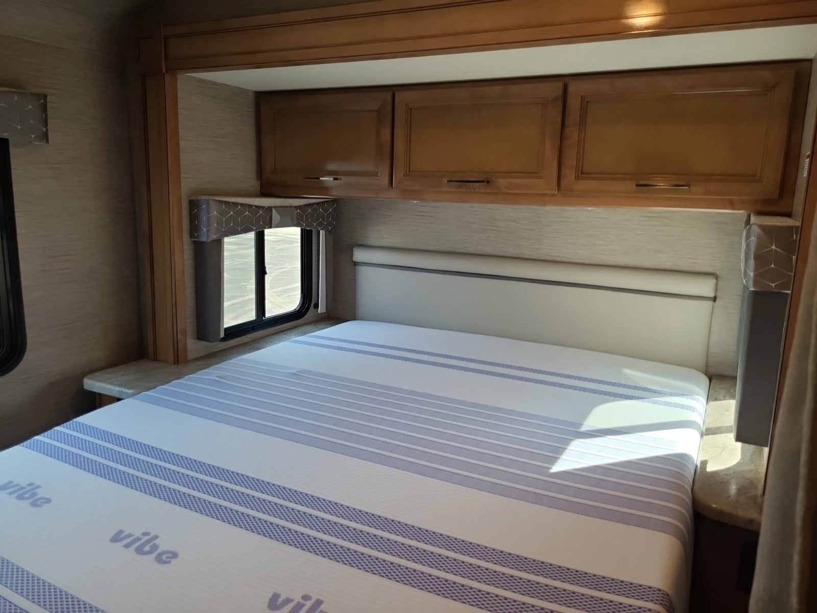 RV rental bedroom: a comfortable bed with striped mattress, light headboard, overhead cabinets, and side windows.. Thor Quantum 2021