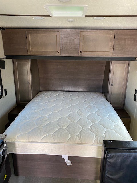 Inside an RV rental: a cozy bedroom with a white quilted queen mattress, overhead cabinets, and side storage.. Sunset Park RV Sun Lite 2021
