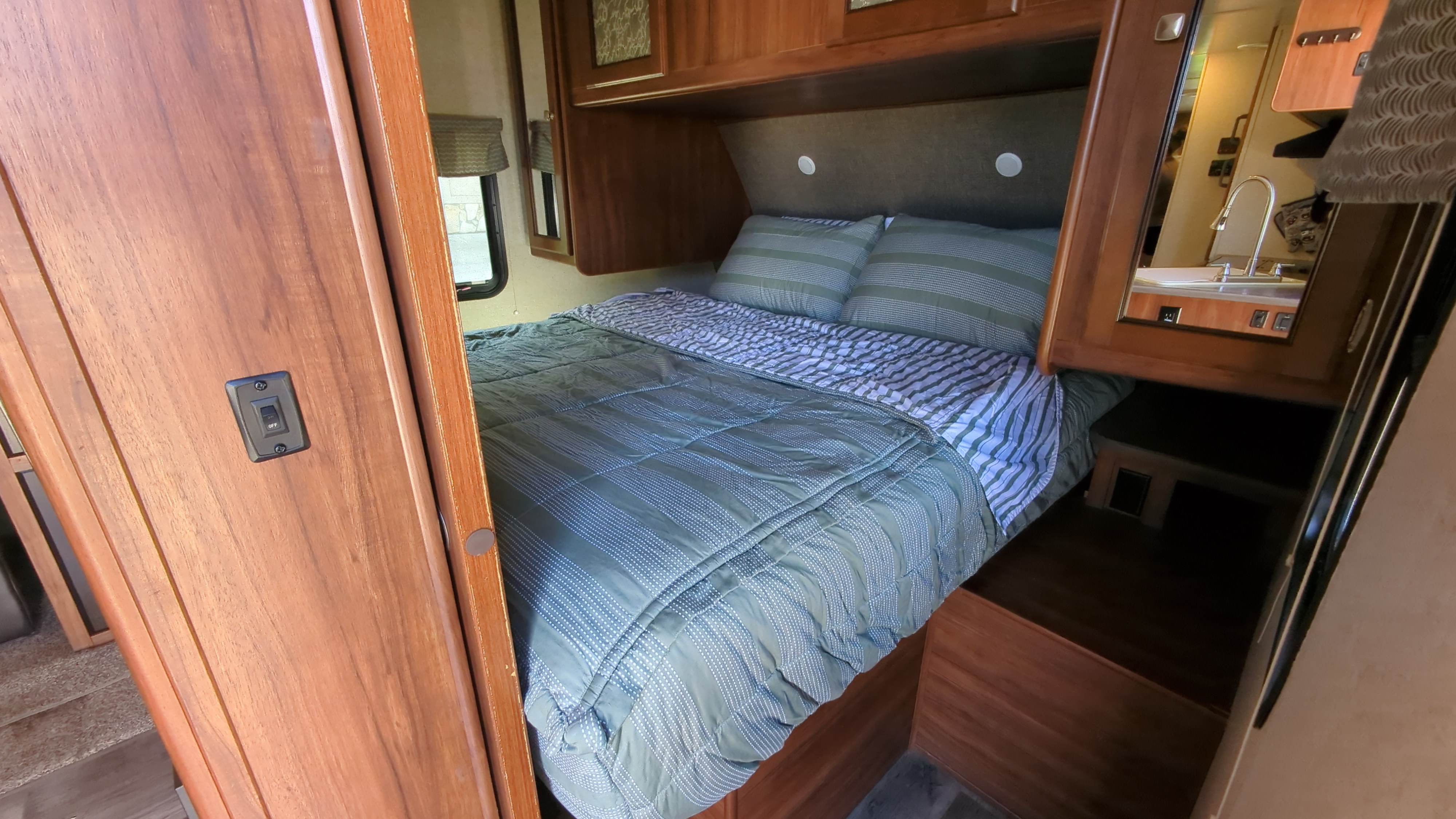 RV rental bedroom view: a cozy bed with striped bedding, wooden cabinetry above, and a mirror reflecting a sink area on the right.. Heartland RVs Mallard 2017