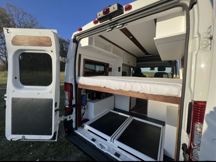Rear view of a white custom camper van RV rental. Doors open reveal a comfortable bed, mattress, and pull-out storage units.. Ram Promaster 2020