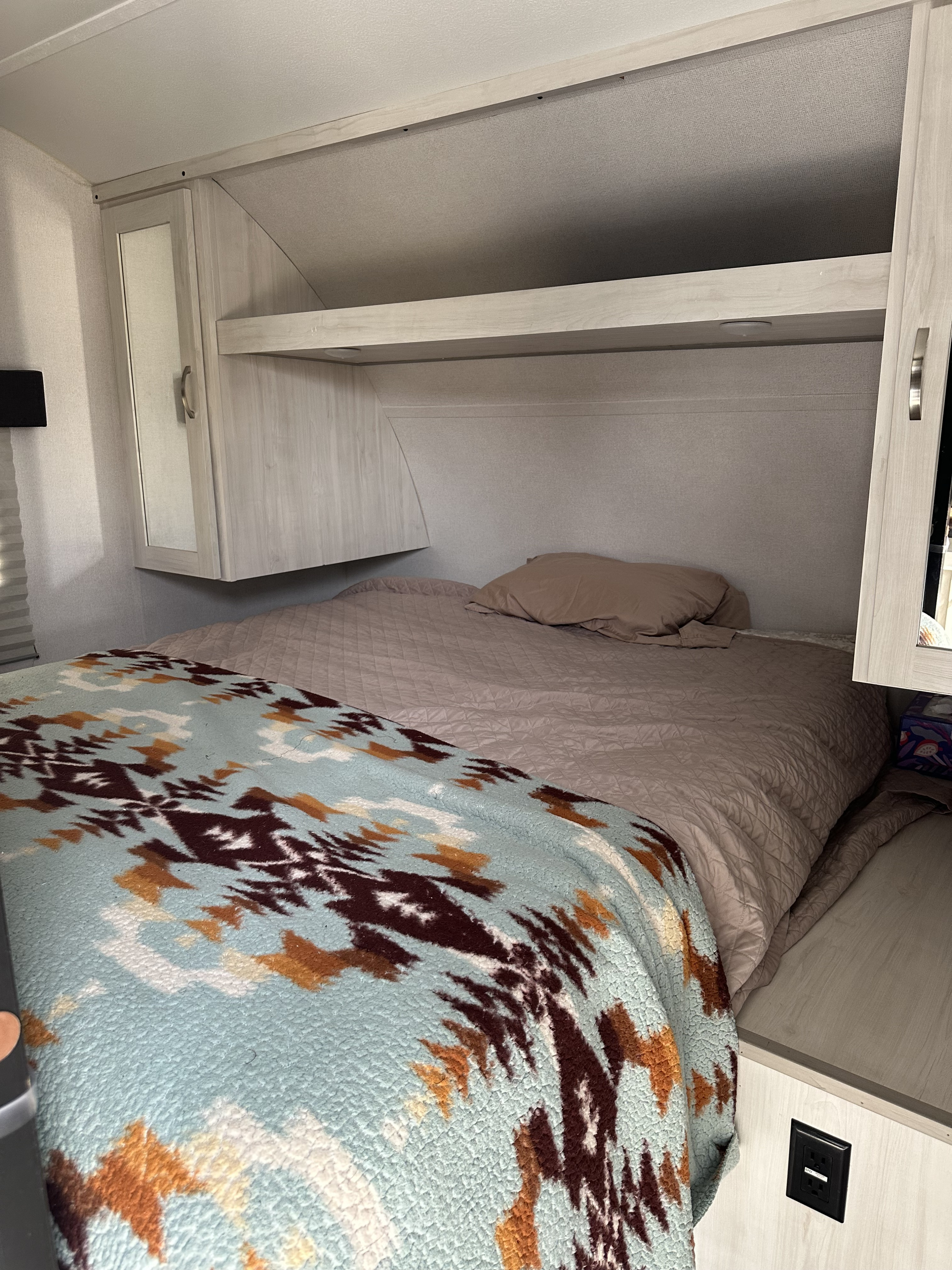 Cozy rv rental bedroom featuring a patterned blanket on the bed, light wood cabinets, shelves, and an electrical outlet.. Forest River Tracer 2021