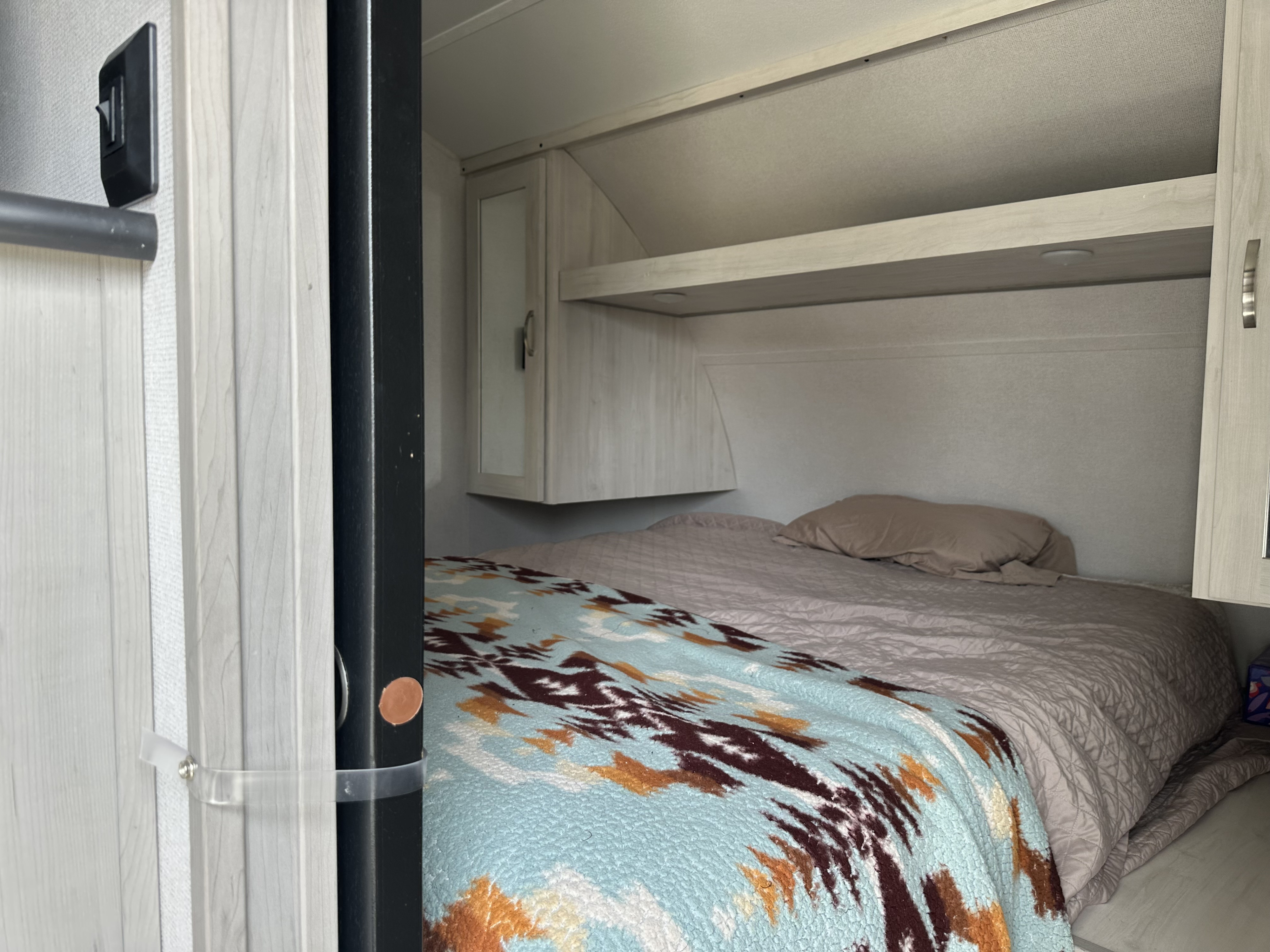 Interior of an RV rental bedroom showing a bed with a patterned blanket, overhead cabinets, and a light switch.. Forest River Tracer 2021