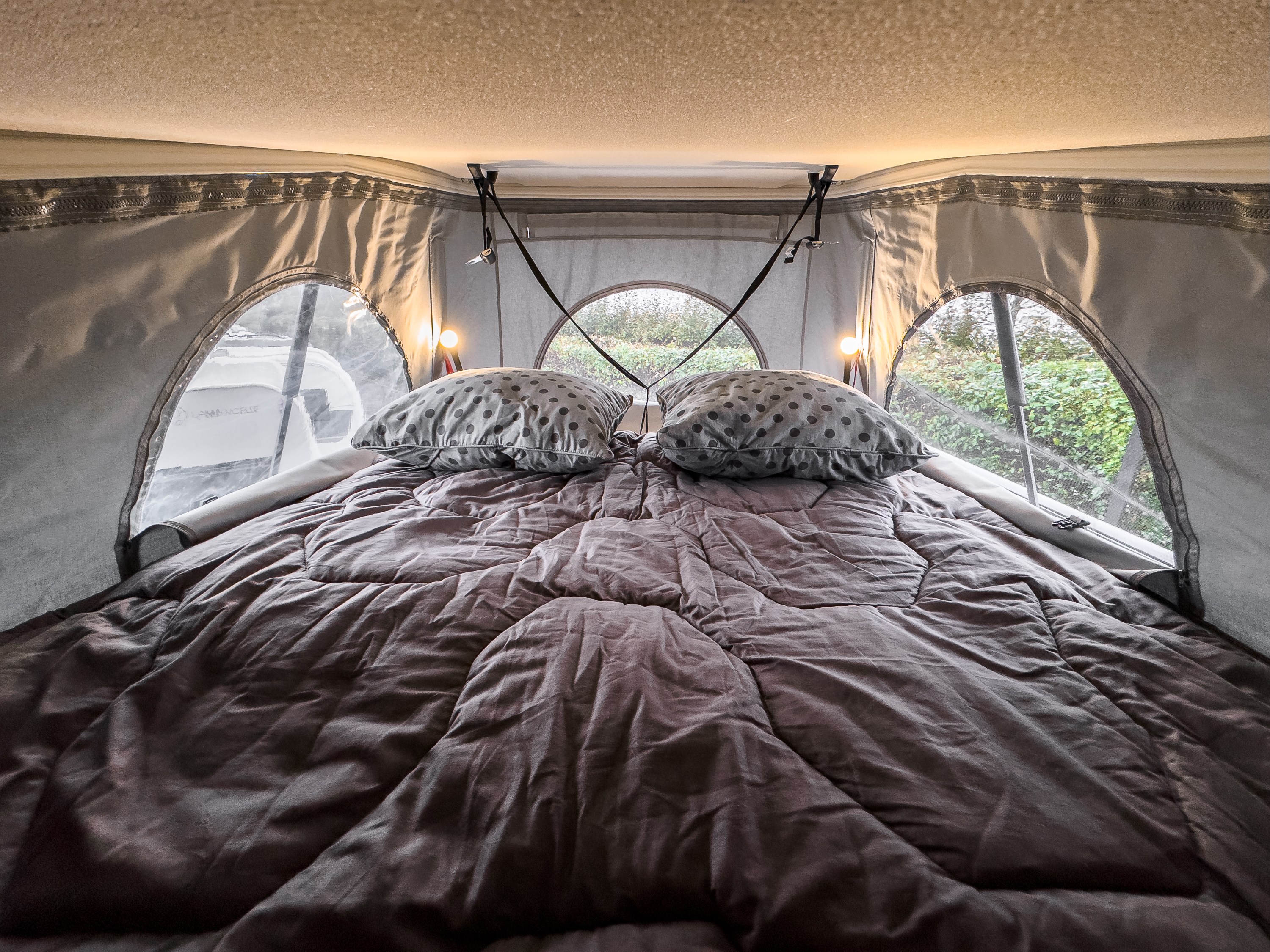 Comfortable bed inside an RV rental with polka dot pillows, warm lights, & window views. Ready for travel camping.. Chausson V594 TOIT RELEVABLE 2025