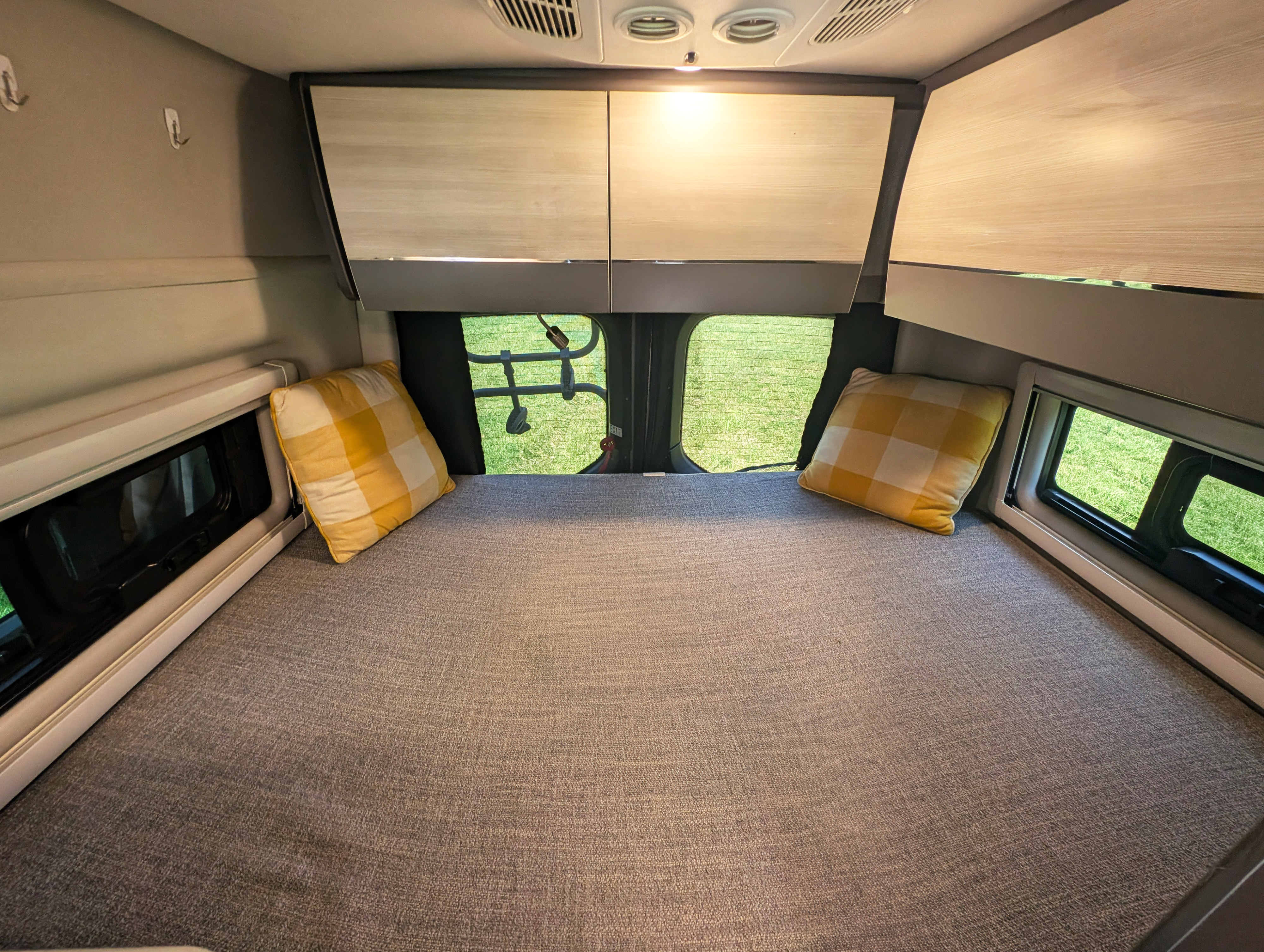 Interior of an **RV rental** showing a neatly made bed with plaid pillows, overhead cabinets, and bright windows with outdoor views.. Thor Sanctuary 2021