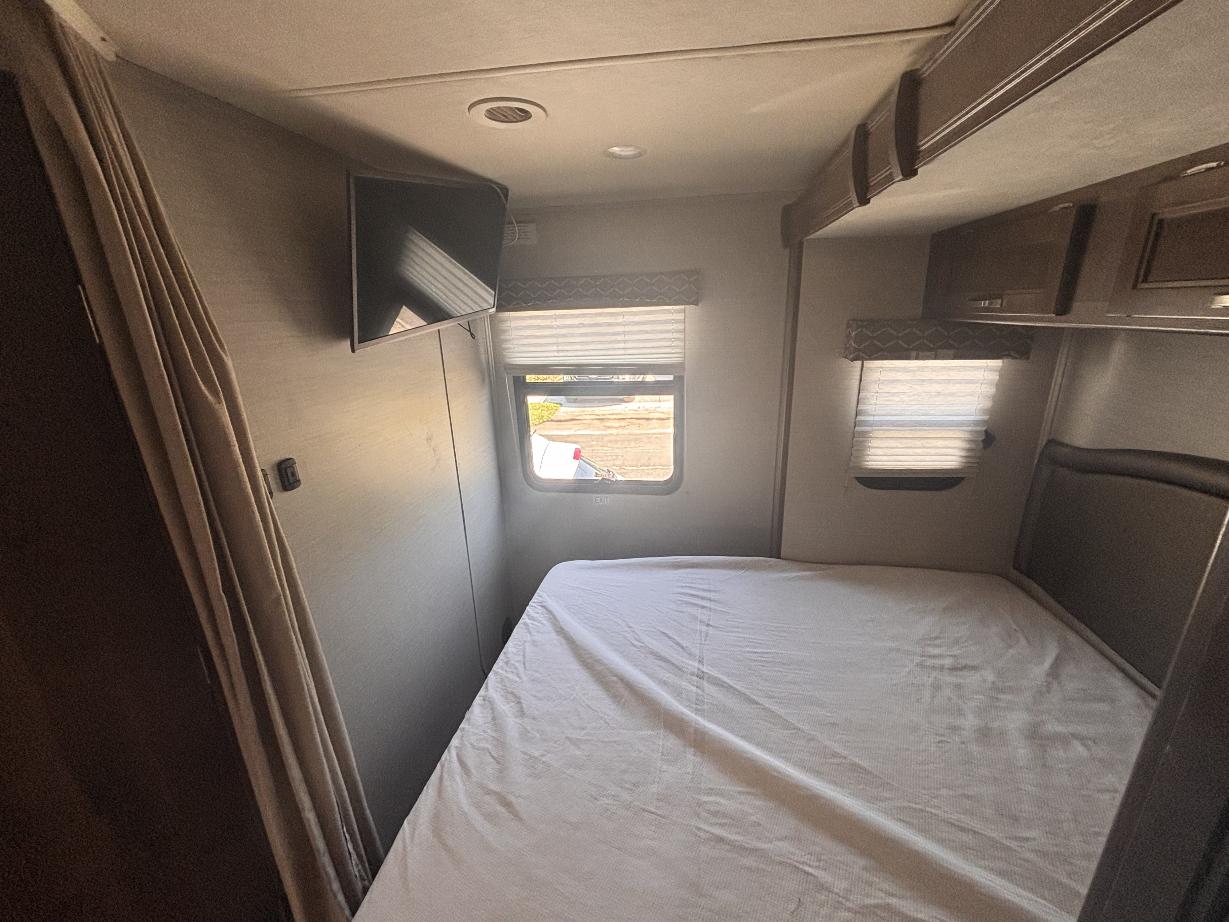 RV rental bedroom featuring a comfortable bed, wall-mounted TV, windows with blinds, and practical overhead storage.. Jayco Melbourne 2020