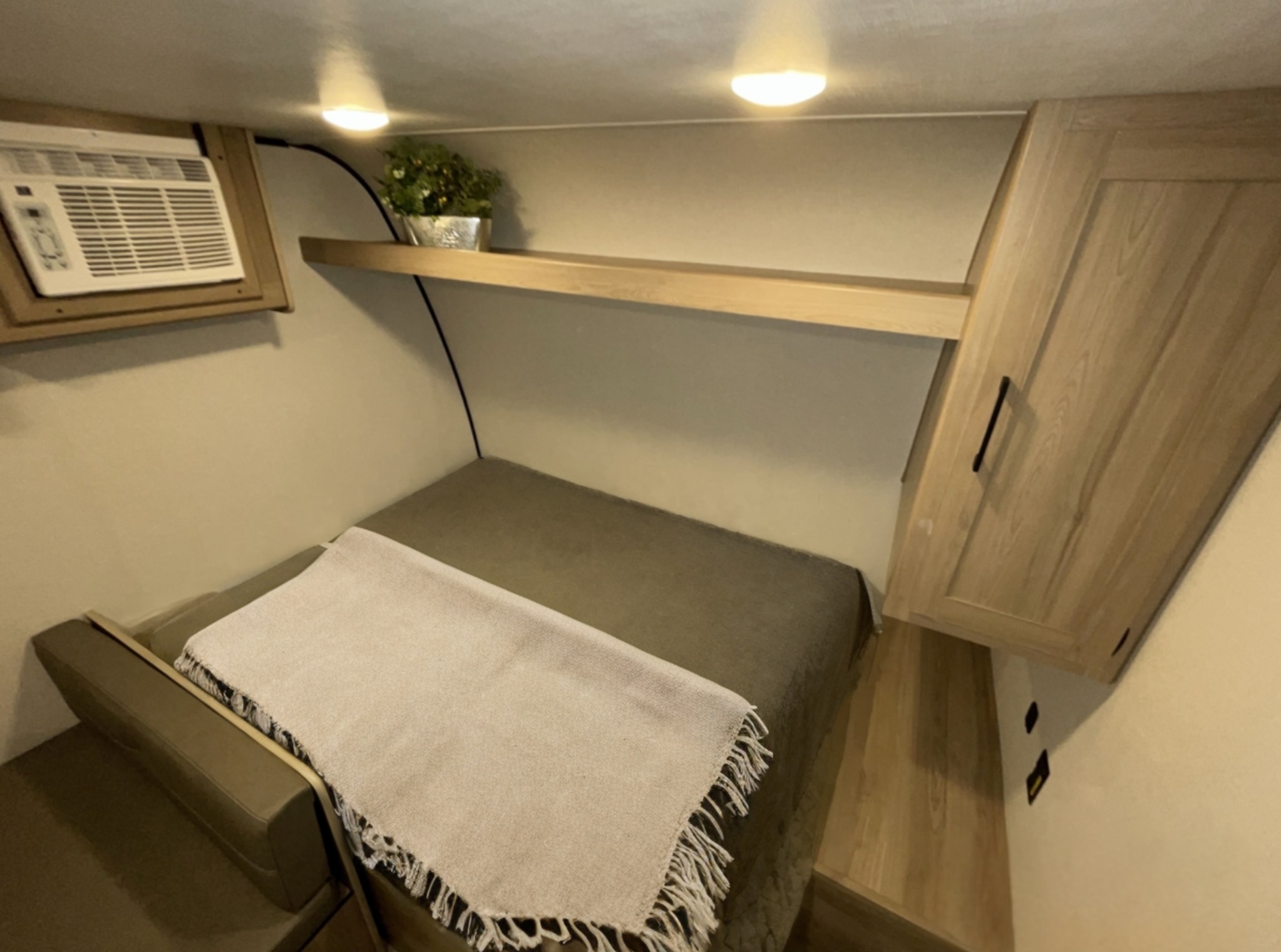 RV rental sleeping area: compact bed, AC, overhead shelf with plant, & wooden cabinet. Cozy space.. Keystone Coleman 2025