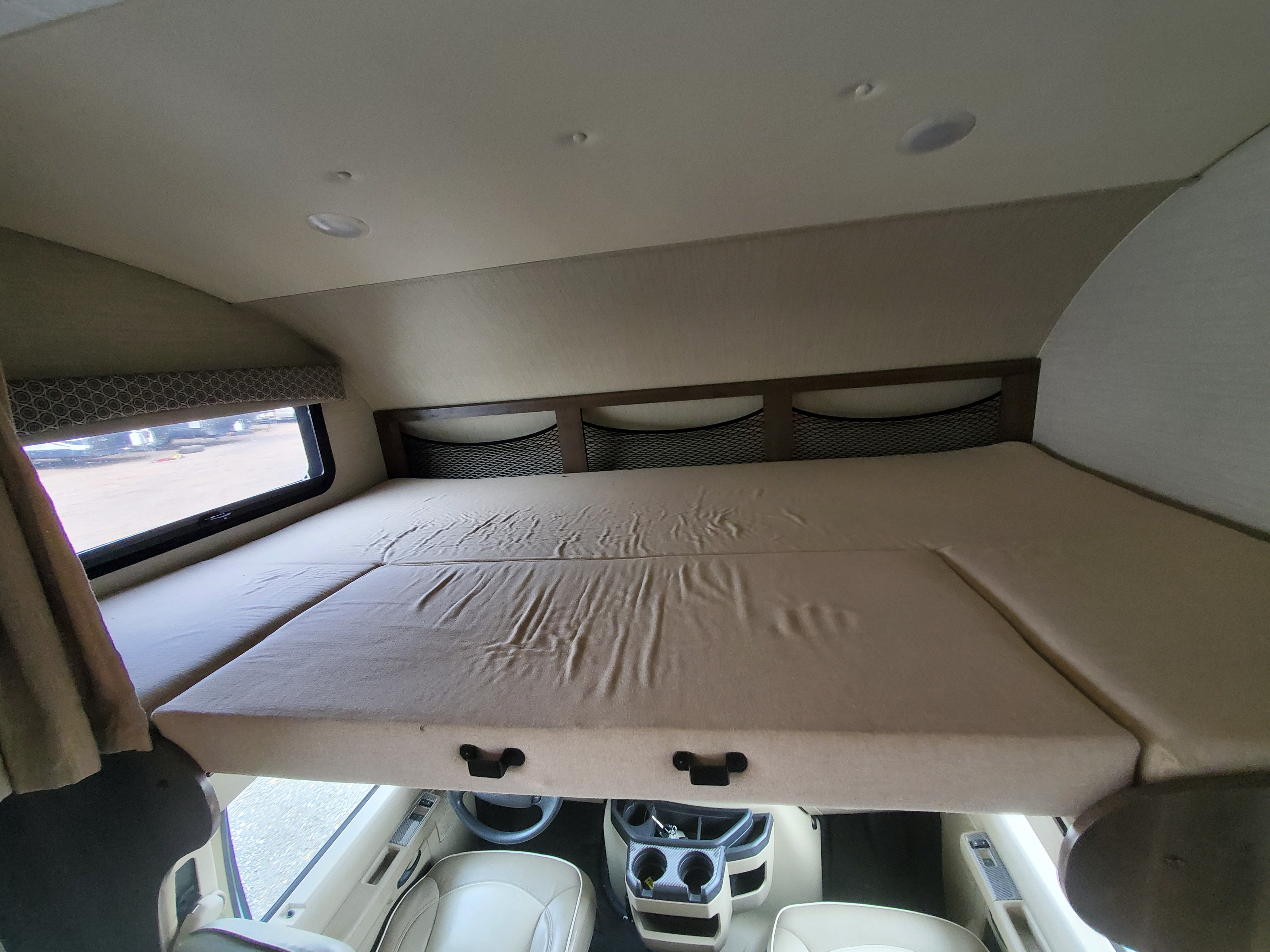 Spacious overhead bunk bed in an RV rental, showing a window, storage nets, and the driver's cab below.. Jayco Greyhawk 2019