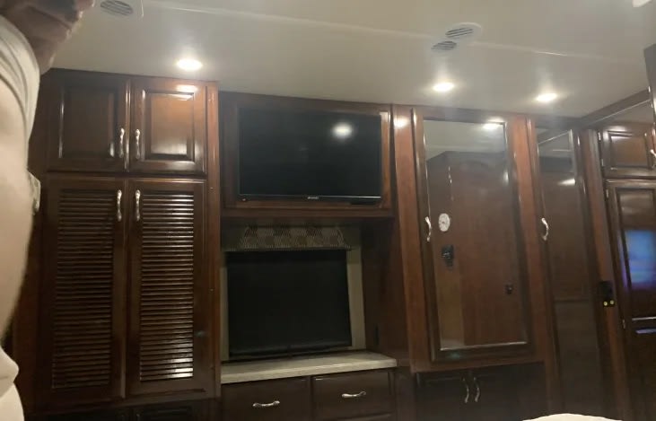 Inside an RV rental: bedroom area with rich wood cabinetry, two TVs, and mirrored wardrobe for ample storage.. Fleetwood storm 32A 2018