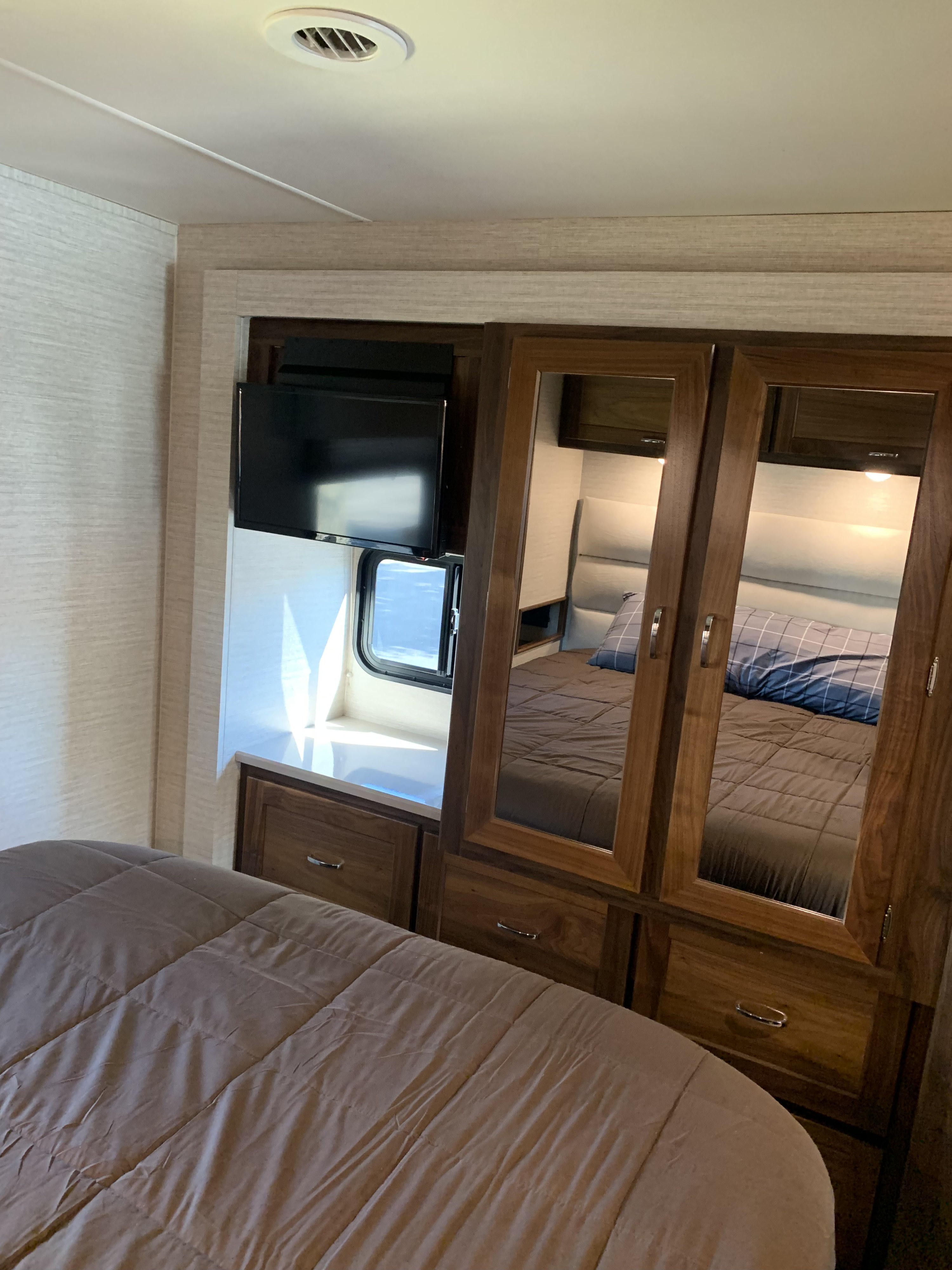 Compact RV rental bedroom featuring a bed with soft comforter, mirrored wardrobe, TV, and window.. Winnebago Intent 2020