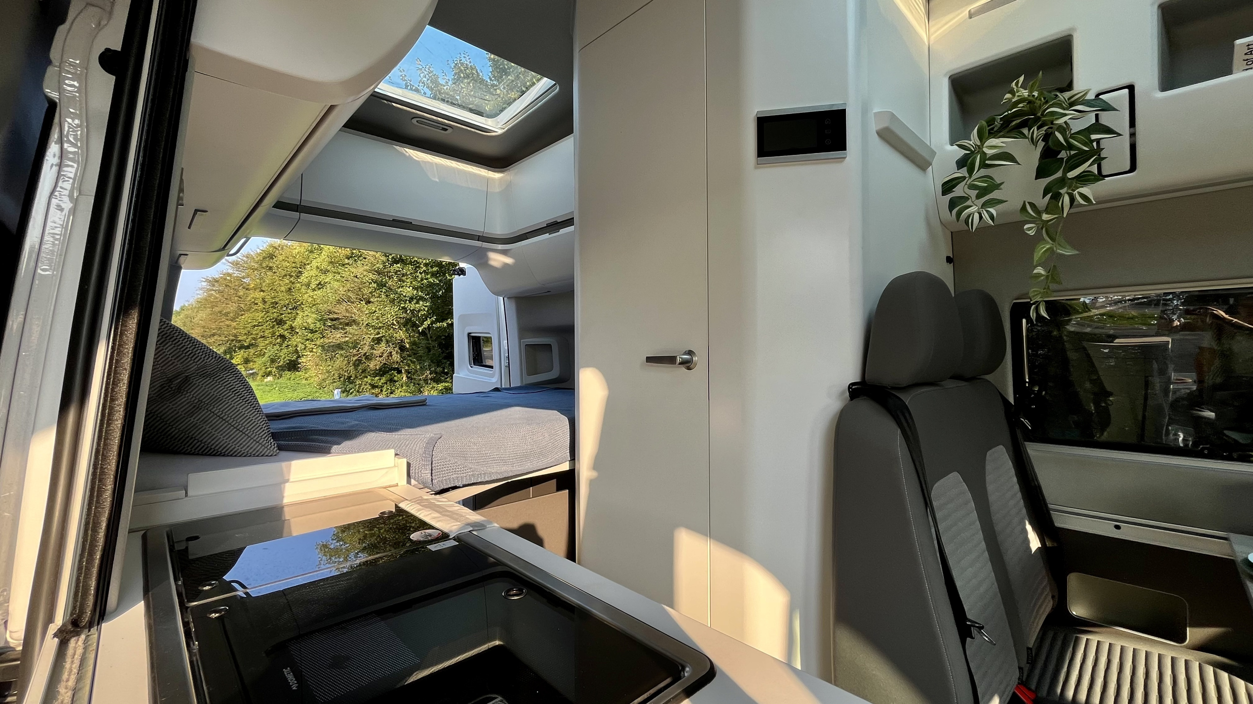 Bright and modern RV rental interior featuring a bed, kitchen, control panel, and roof window with trees outside.. Volkswagen Grand California 2022