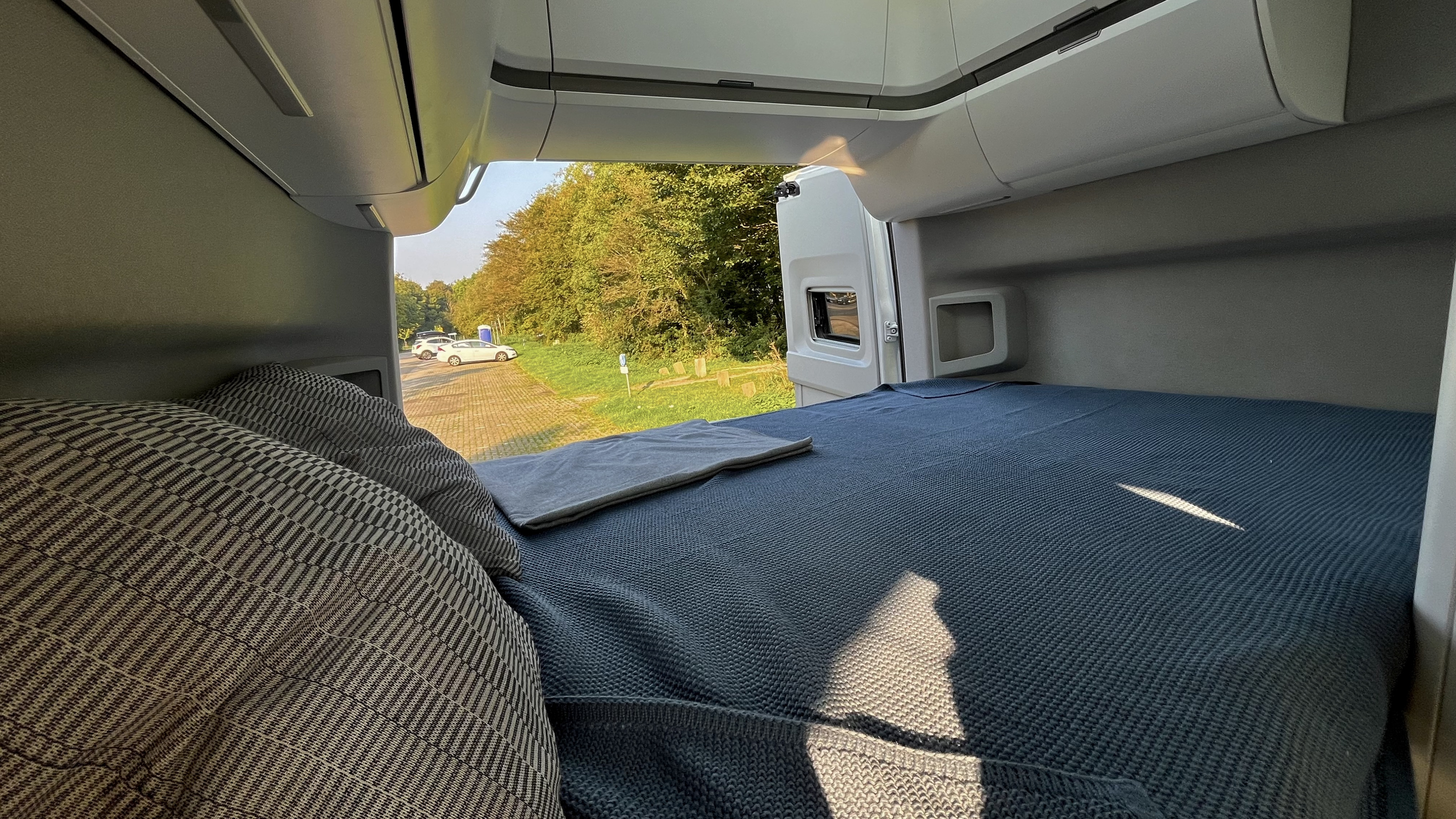 Inside a modern **rv rental**: a comfy bed with blue covers and pillows, offering views of a sunny parking area and trees.. Volkswagen Grand California 2022