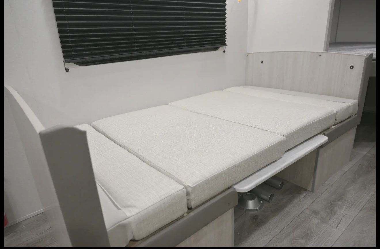 RV rental interior: A beige convertible bed/seating with light wood accents and a pleated window blind, offering compact comfort.. Coachmen Catalina 2025