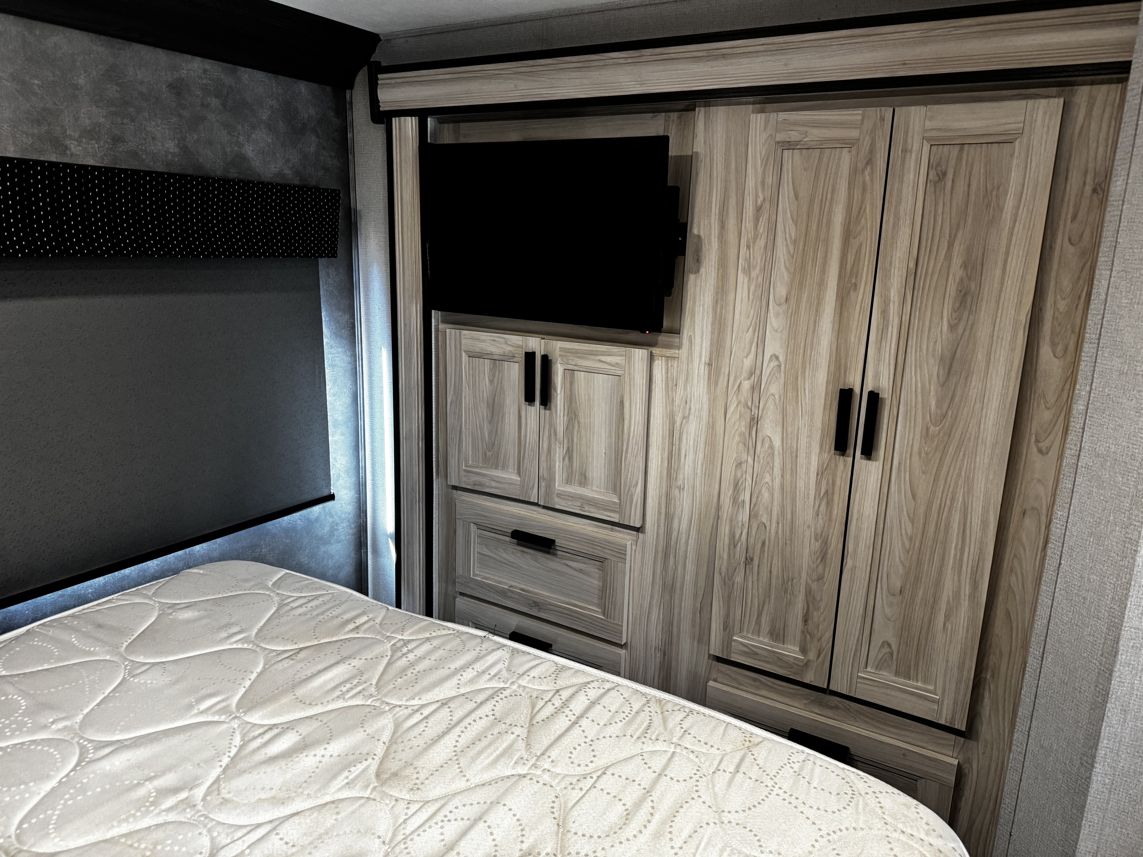 This RV rental bedroom features a white mattress, built-in wooden cabinets, a mounted TV, and textured dark gray walls.. Forest River Forester 2022