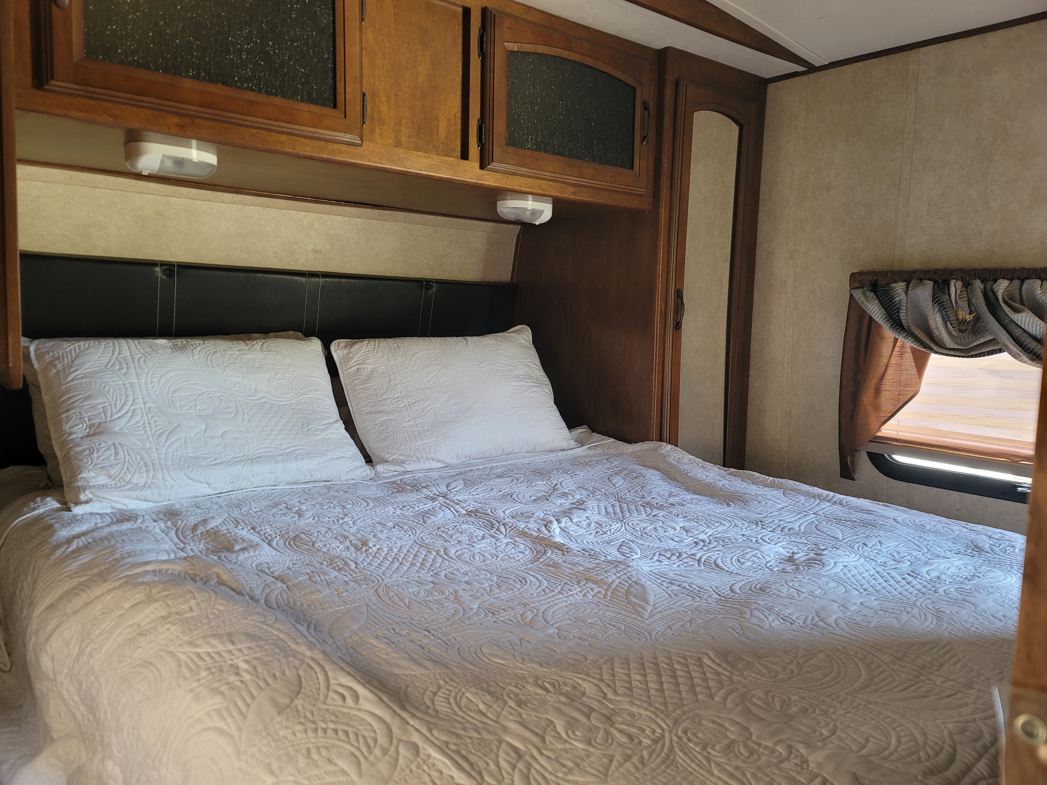 RV rental bedroom with a white quilted bed, black headboard, overhead wooden cabinets, and a window.. Forest River Wildwood Heritage Glen Lite 2014