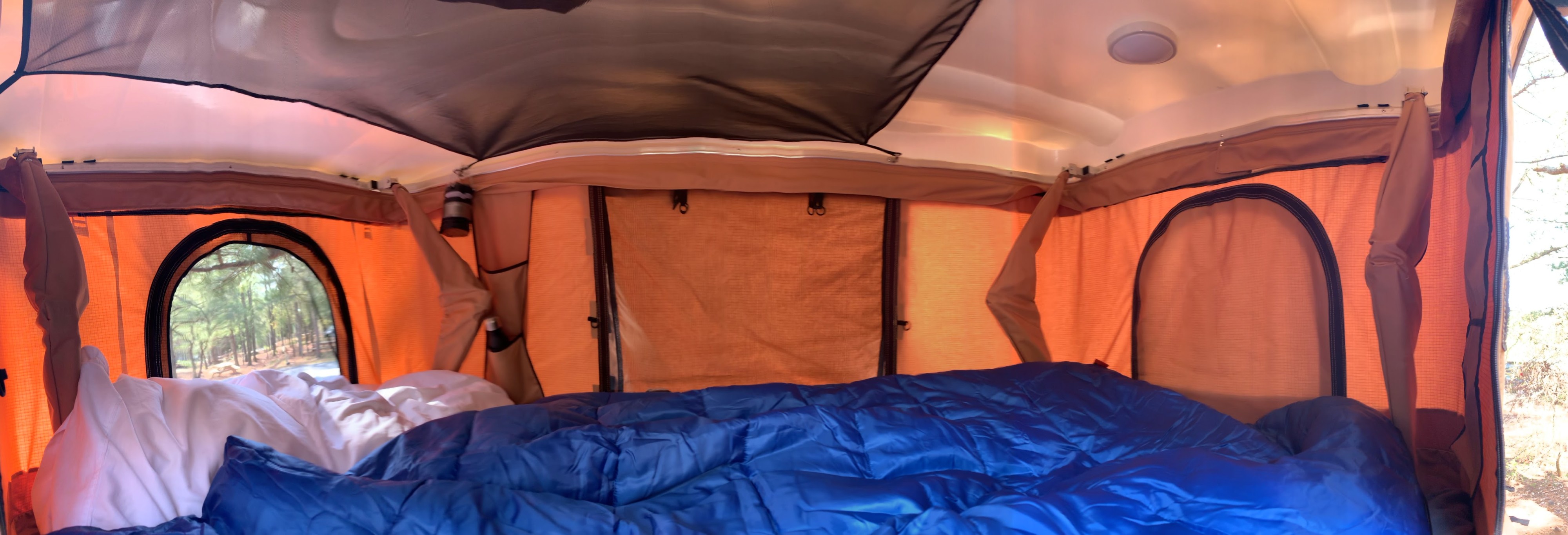 Cozy RV rental interior with a comfortable bed, surrounded by windows offering a peaceful forest view, perfect for camping.. Jeep Renegade 2020