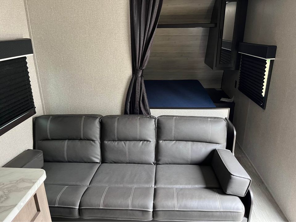 This RV rental living area features a convertible sofa, bunk bed, and windows with pleated blinds, offering a compact travel space.. Jayco Jay Flight SLX 2022