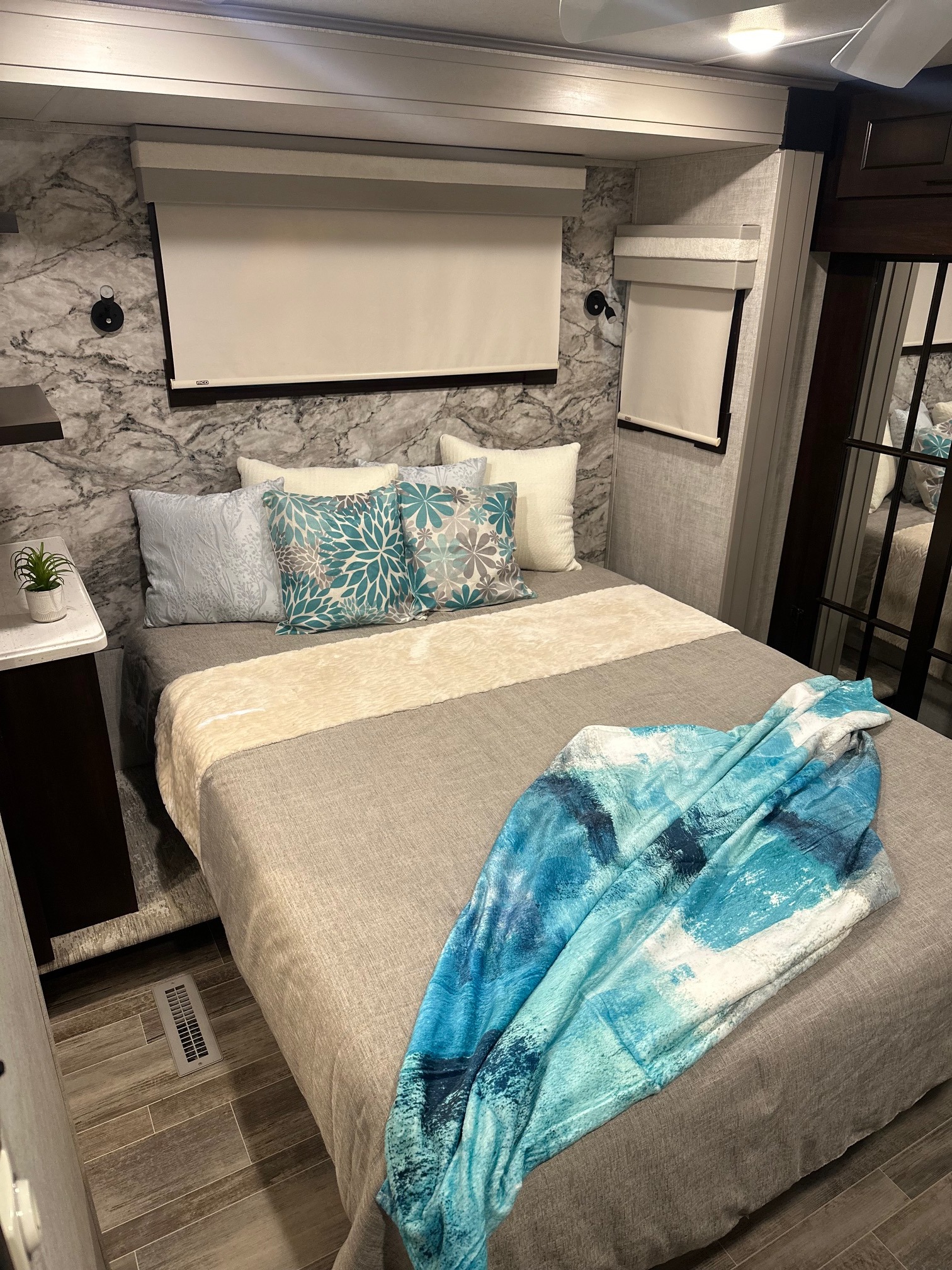 Modern RV rental bedroom featuring a bed with patterned pillows, grey bedding, and a vibrant blue throw, along with wood-look floors.. Forest River Palomino 2021