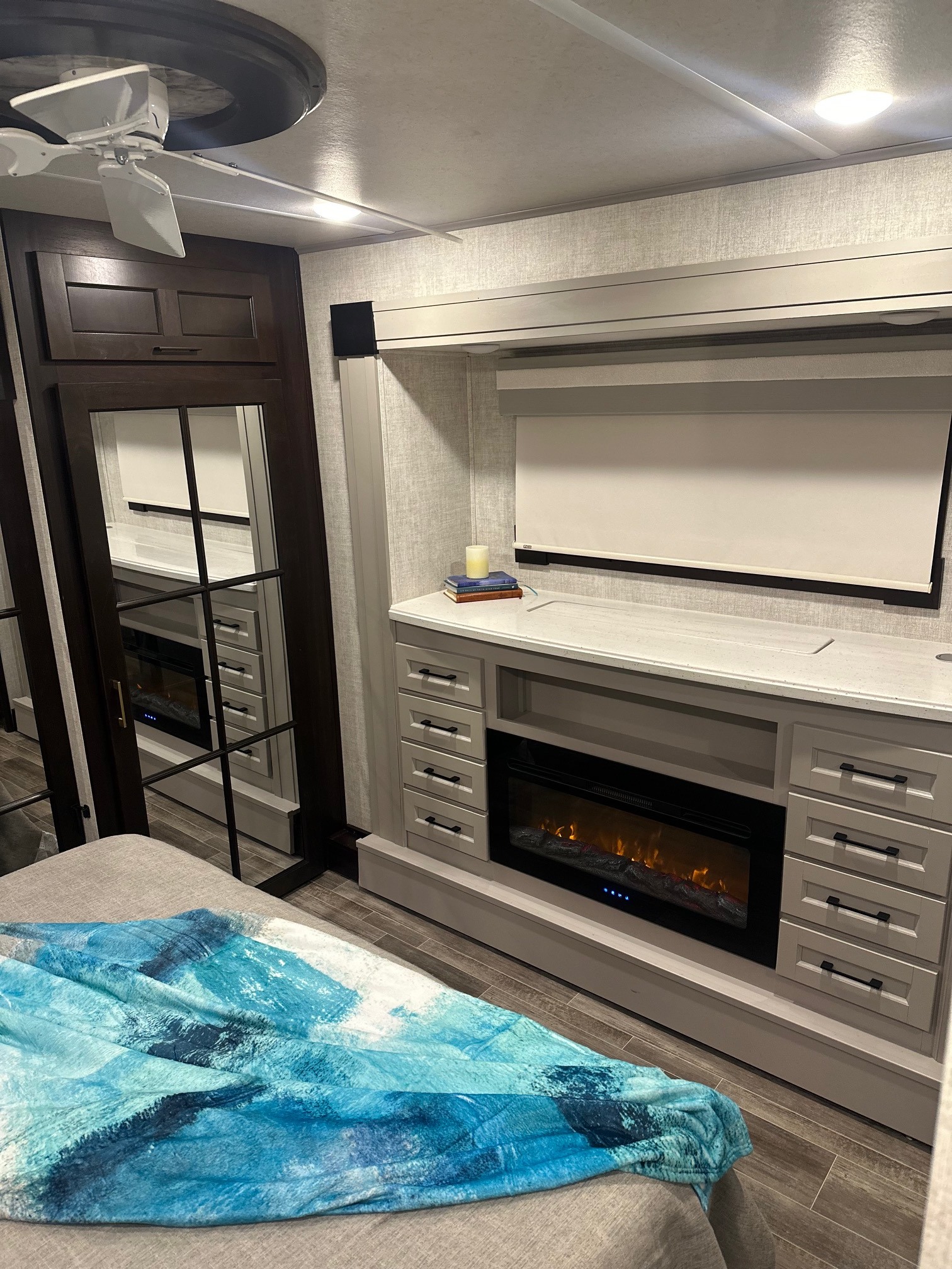 Modern RV rental bedroom. Features a fireplace, ceiling fan, roller blind, mirrored door, and a cozy bed with a blue blanket.. Forest River Palomino 2021