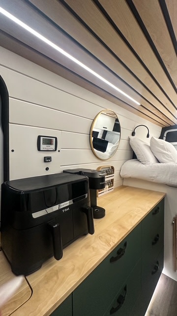 Inside a modern RV rental: kitchen setup with T-fal air fryer, Keurig, wood counters, green cabinets, and a cozy bed area.. Mercedes-Benz Sprinter 2021