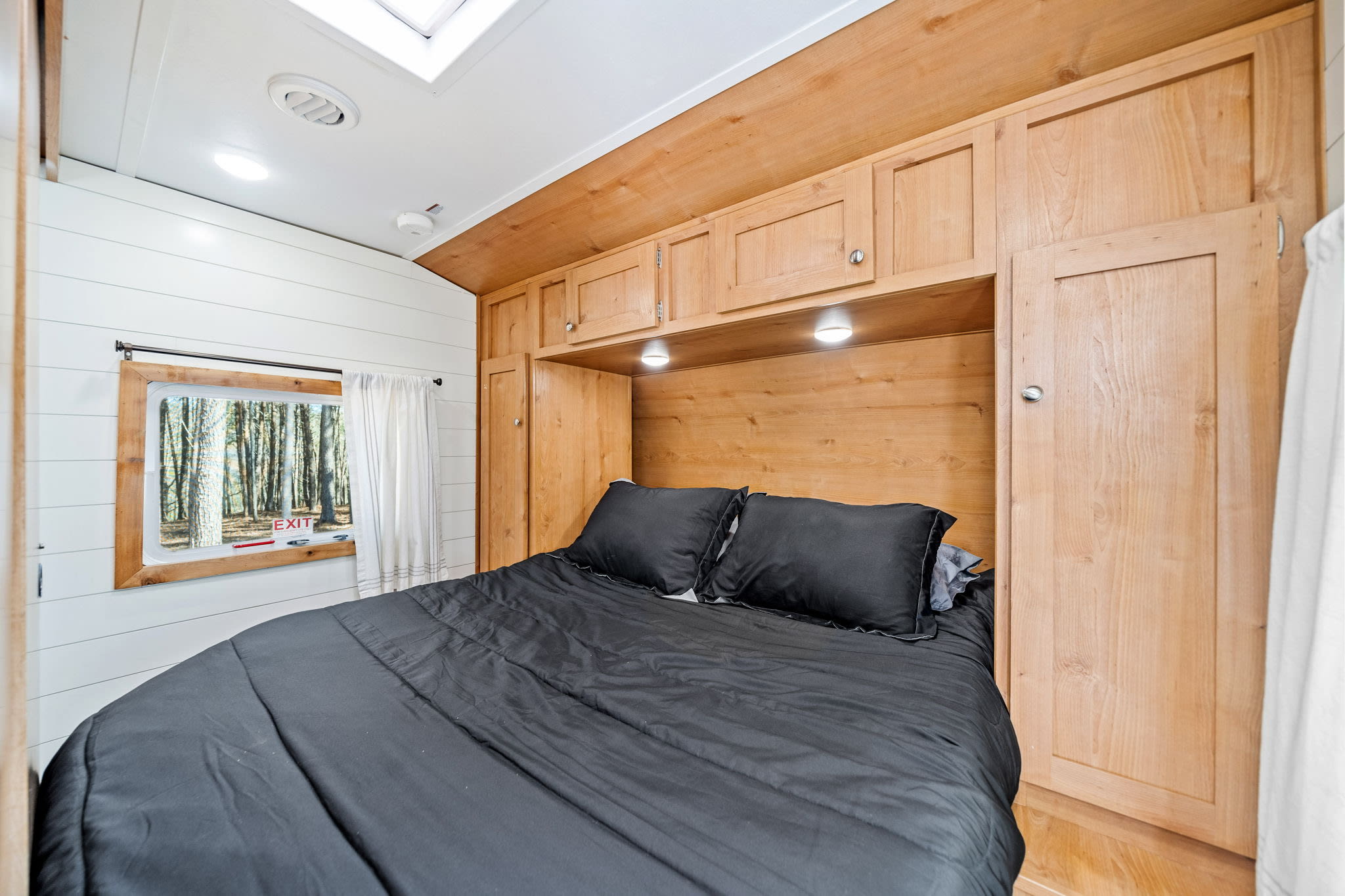 Cozy RV rental bedroom with dark bedding, built-in wooden cabinets, and a window overlooking a forest.. Riverside RV Retro 2020
