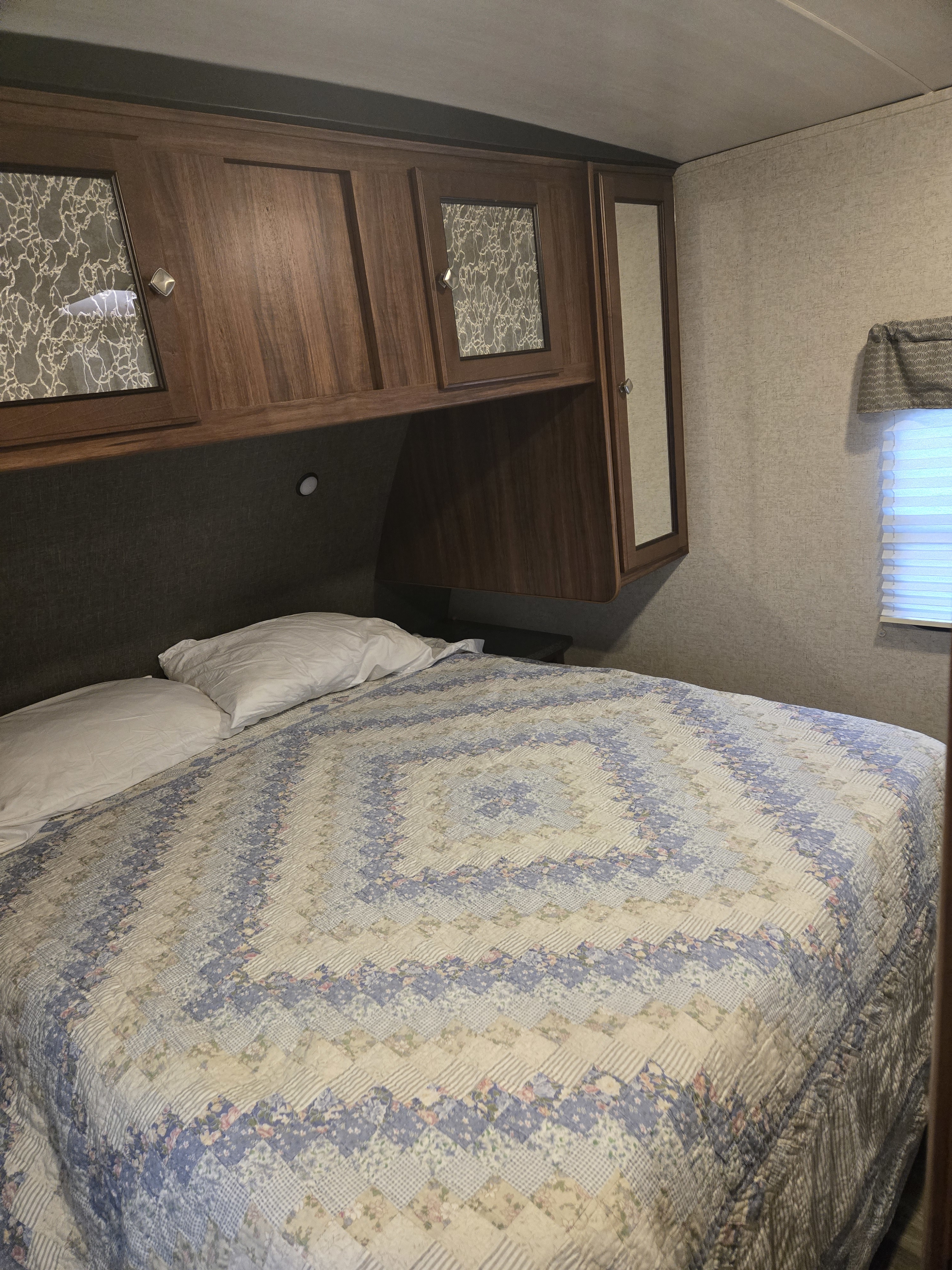 Cozy rv rental bedroom features a bed with a colorful quilt, overhead wood cabinets, and window blinds, ideal for travel.. Heartland RVs Wilderness 2017