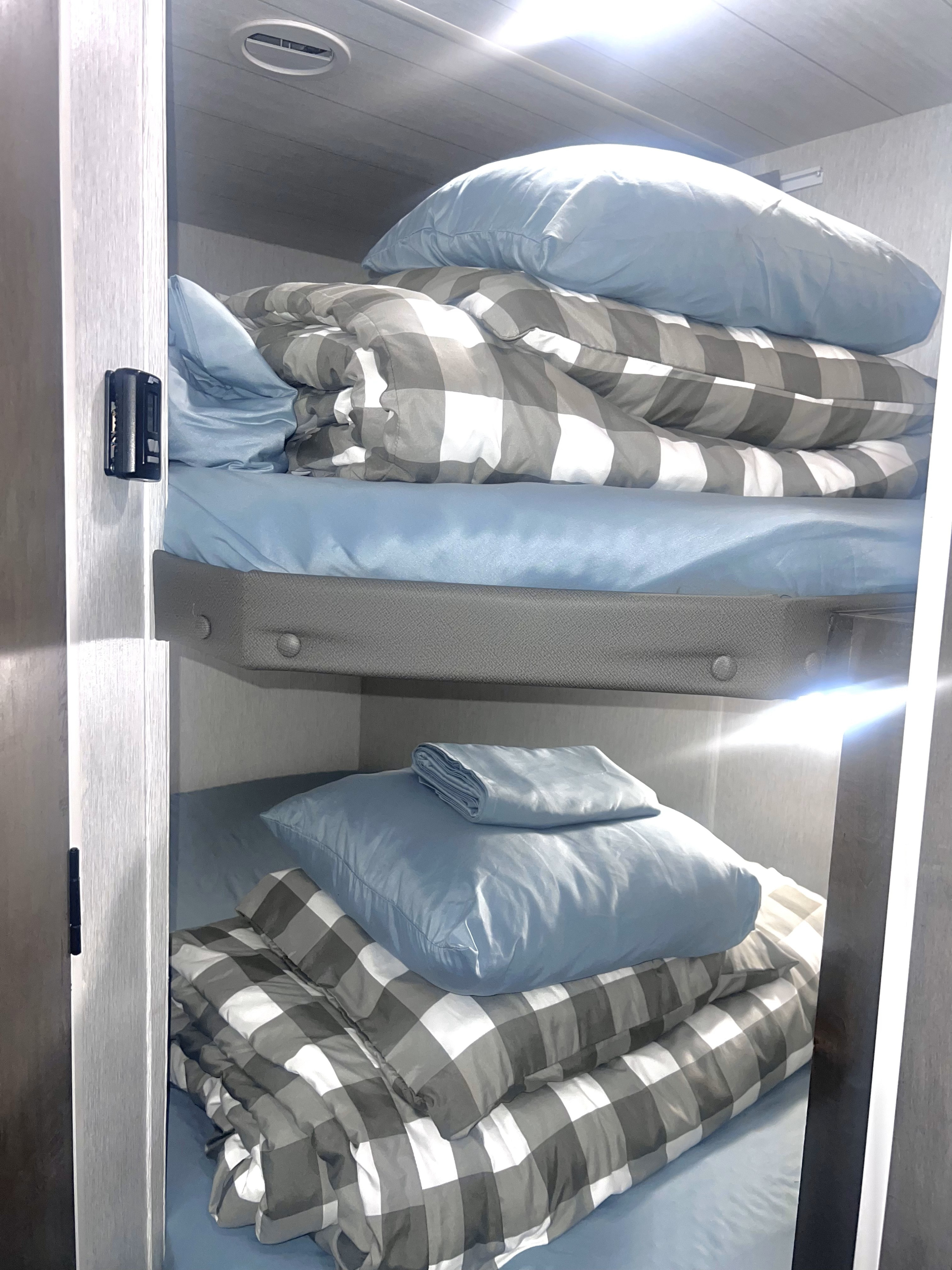 Inside an RV rental: two well-made bunk beds. Light blue bedding and cozy grey & white checkered comforters, perfect for travelers.. Forest River Wildwood X-Lite 2022