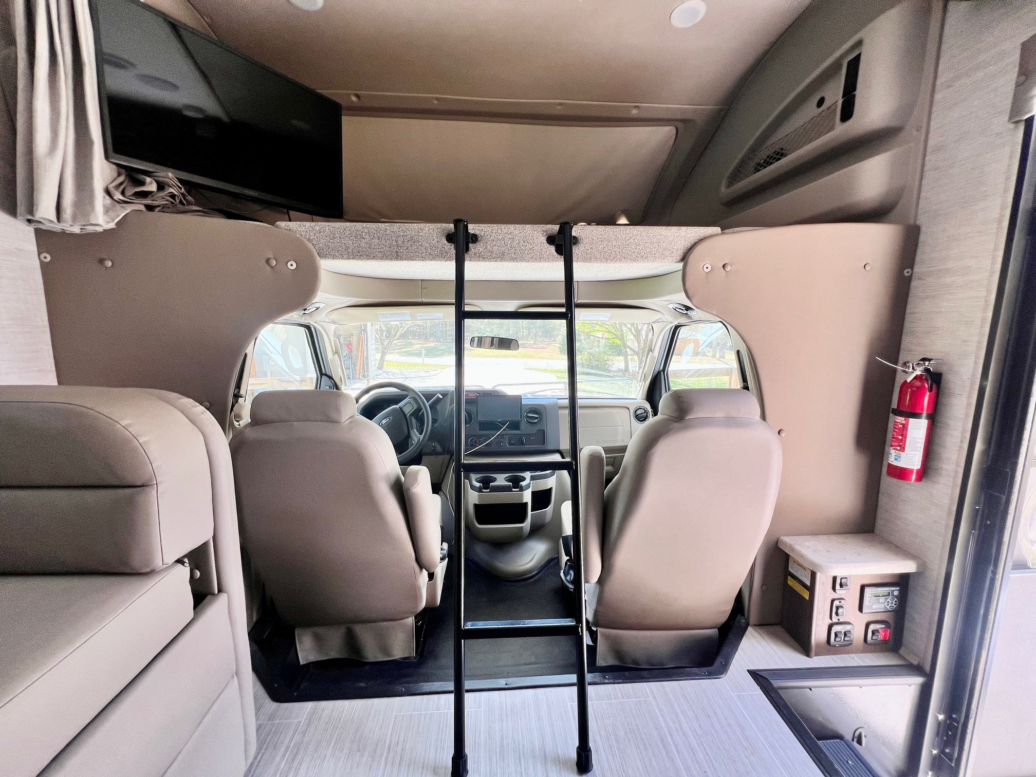 Inside an RV rental: view of the front cockpit, ladder to the over-cab bunk, TV, controls, and fire extinguisher.. Entegra Coach Esteem 2021