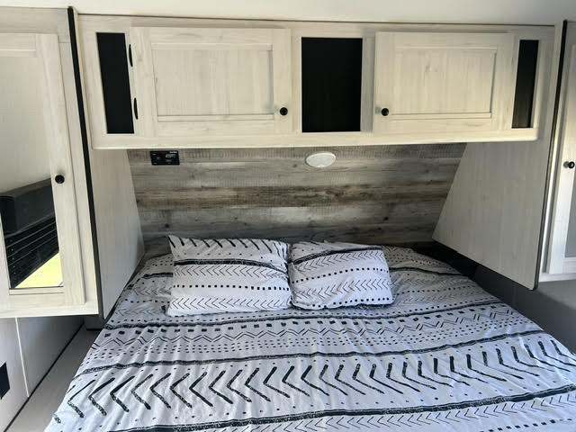This RV rental bedroom features a comfy bed with patterned bedding, overhead storage cabinets, and a rustic wood-look accent wall.. Heartland Lithium 2021