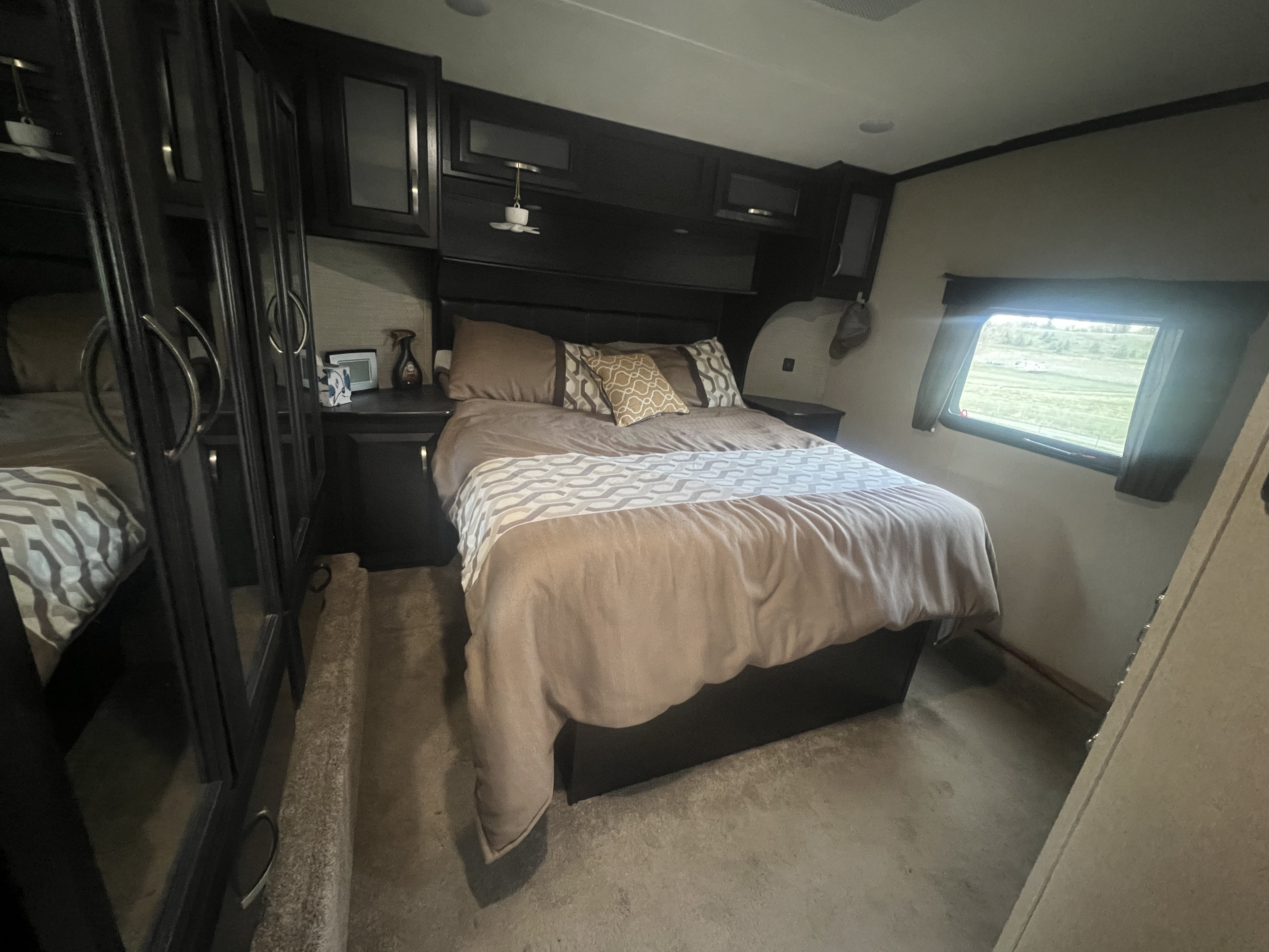 A cozy RV rental bedroom with dark wood built-ins, a comfortable bed, and a bright window overlooking green fields.. Dutchmen Voltage 3305 2018