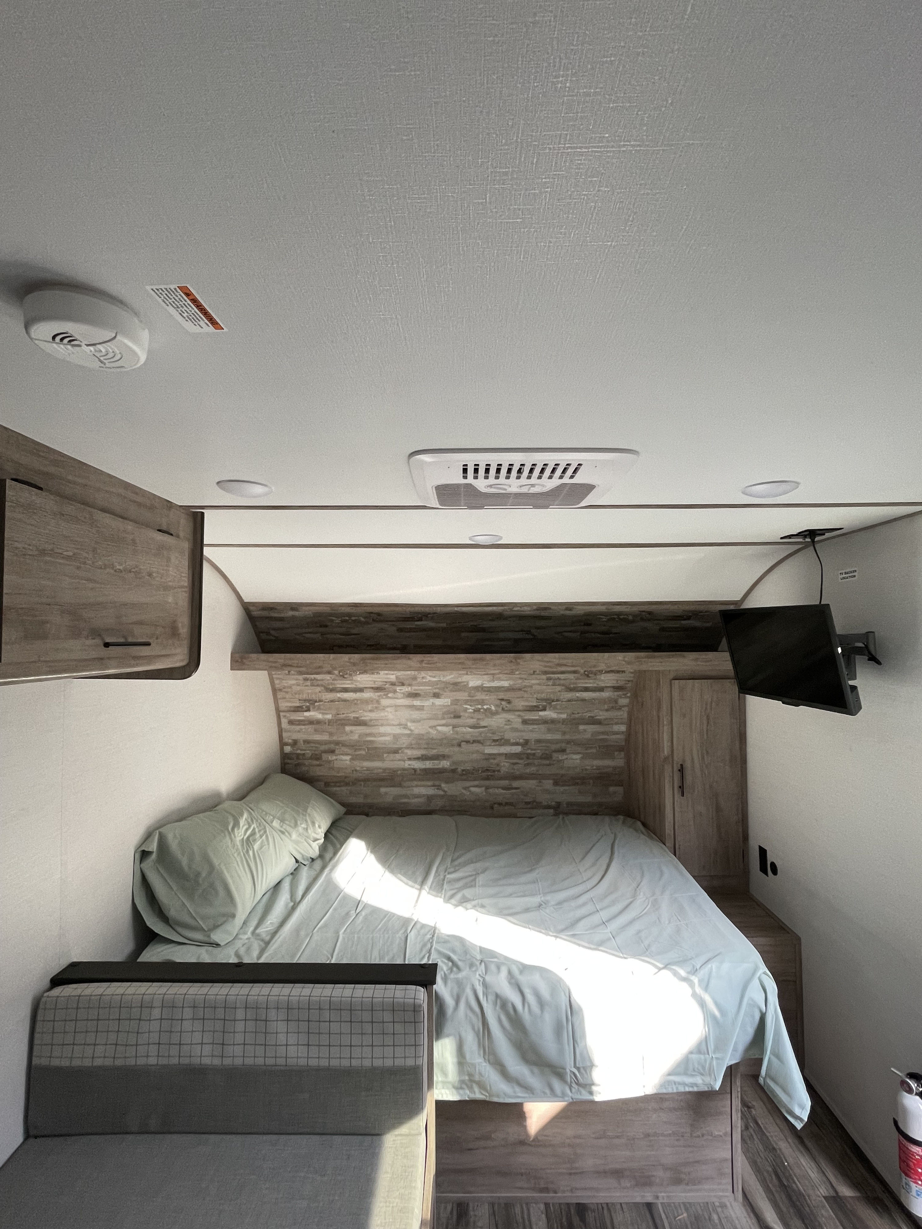 Clean RV rental bedroom featuring a bed, couch, wood cabinets, ceiling AC, TV, smoke detector, and fire extinguisher for safety.. Gulf Stream Ameri-Lite 2024