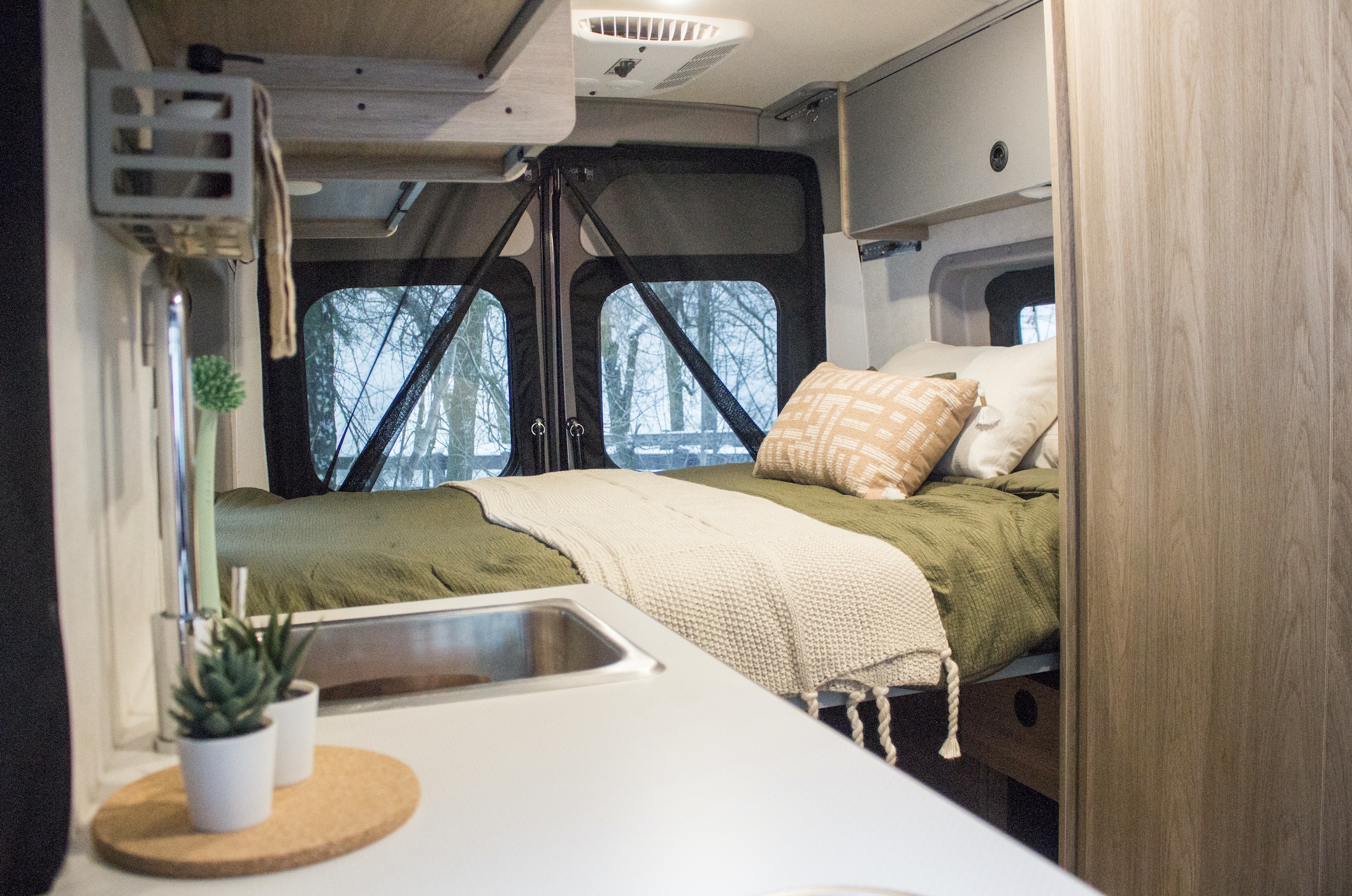 Inside a modern RV rental: a cozy bed with olive green bedding, pillows, and a kitchenette area in a spacious camper van.. Winnebago Solis 2023