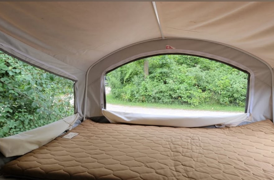 **RV rental** pop-up camper interior: Quilted bed faces large windows with a view of green trees & dirt path.. Forest River Hybrid 16RB 2019