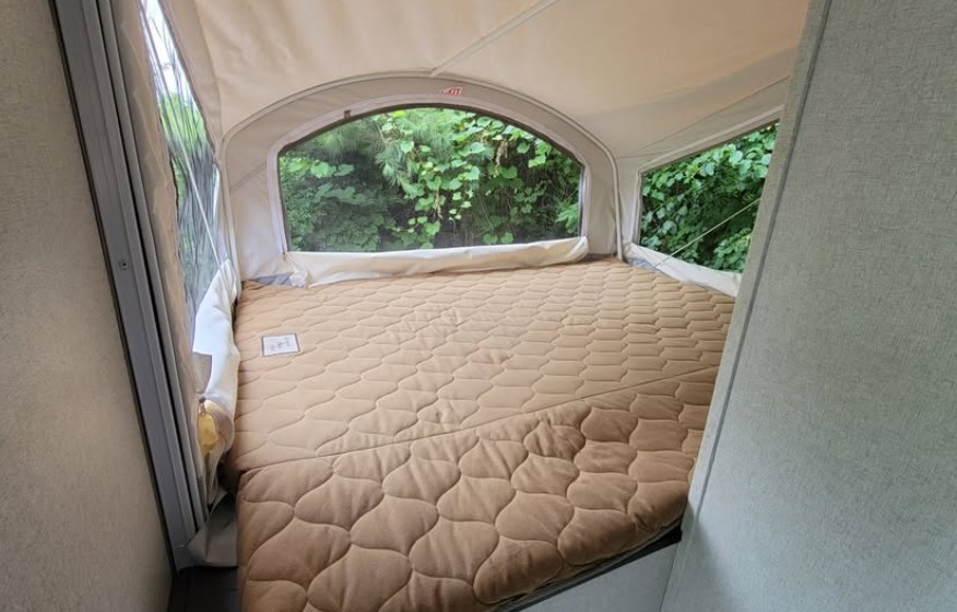 Interior of an RV rental pop-up camper, featuring a large bed with a quilted mattress and windows overlooking green trees.. Forest River Hybrid 16RB 2019
