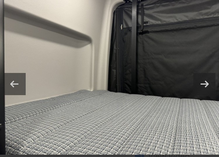 Inside an RV rental: a grey patterned mattress in a compact sleeping area with black fabric walls, ready for your adventure.. Winnebago Revel 2024