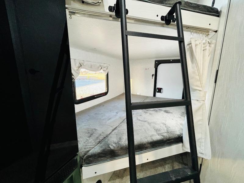 Inside an RV rental: a black ladder ascends to bunk beds. The lower bed, with a gray blanket, is beside a window showing outside.. Forest River No Boundaries NoBo 20.3 2025