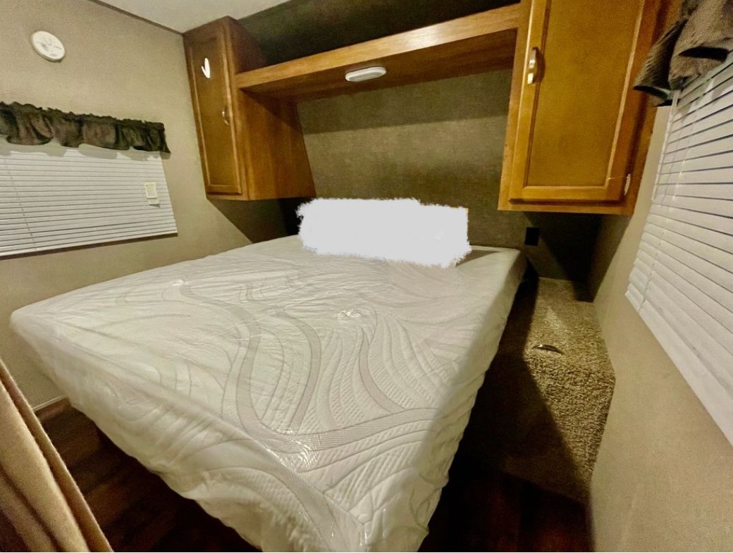 RV rental bedroom with new white mattress in plastic, light wood overhead cabinets, and a window with blinds.. Keystone RV Summerland 2017