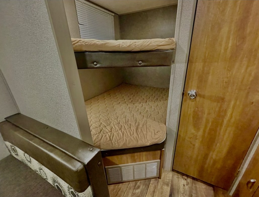 RV rental interior: Compact bunk beds with quilted mattresses are nestled next to a window and wooden door, offering cozy sleeping.. Keystone RV Summerland 2017