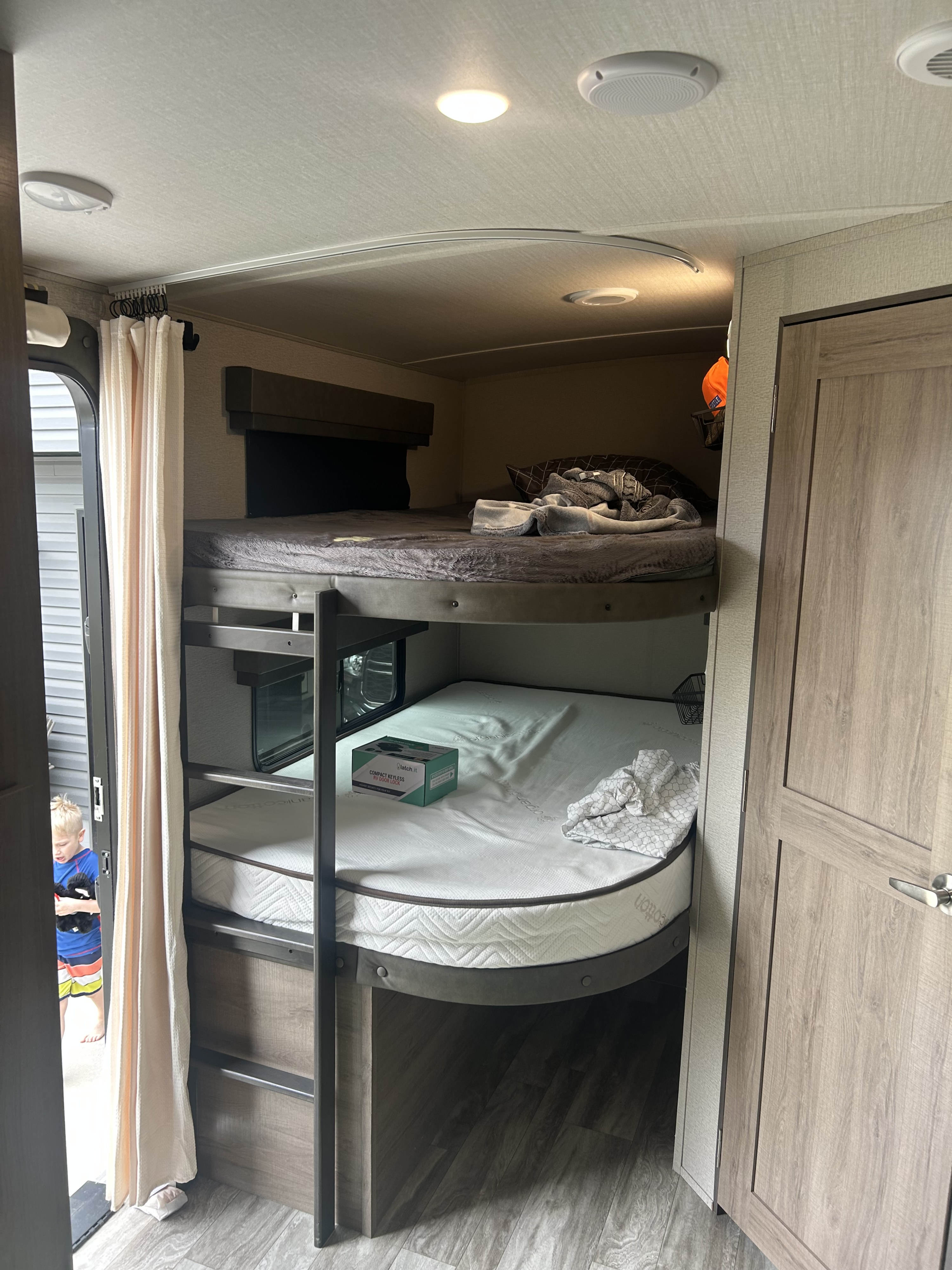 An RV rental interior with comfortable bunk beds. The top bunk has bedding, and the lower has a mattress. A child is by the door.. Grand Design Imagine 2022