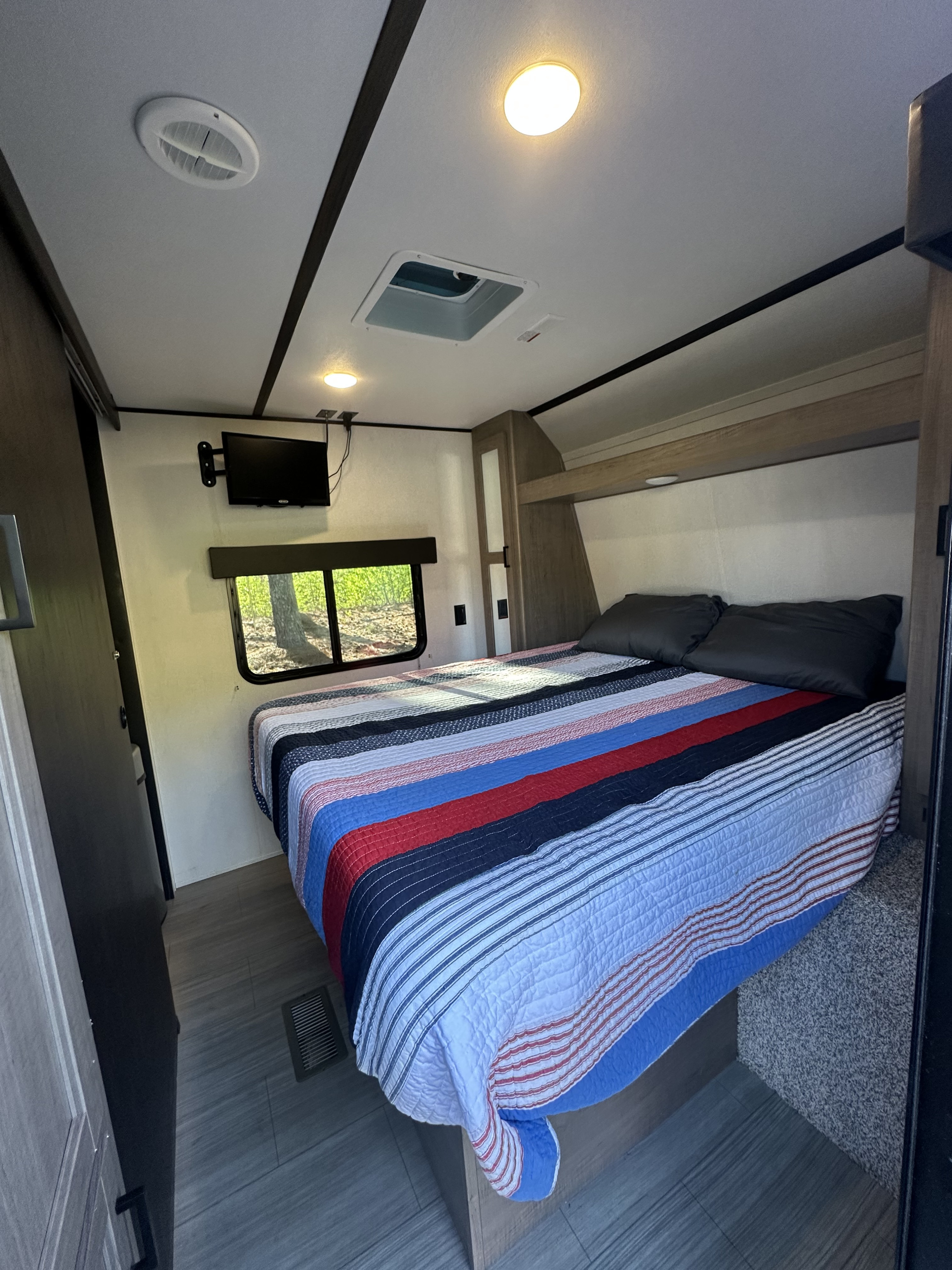 Cozy RV rental bedroom with striped bed, wall-mounted TV, and a window offering natural light.. Keystone Hideout 2021