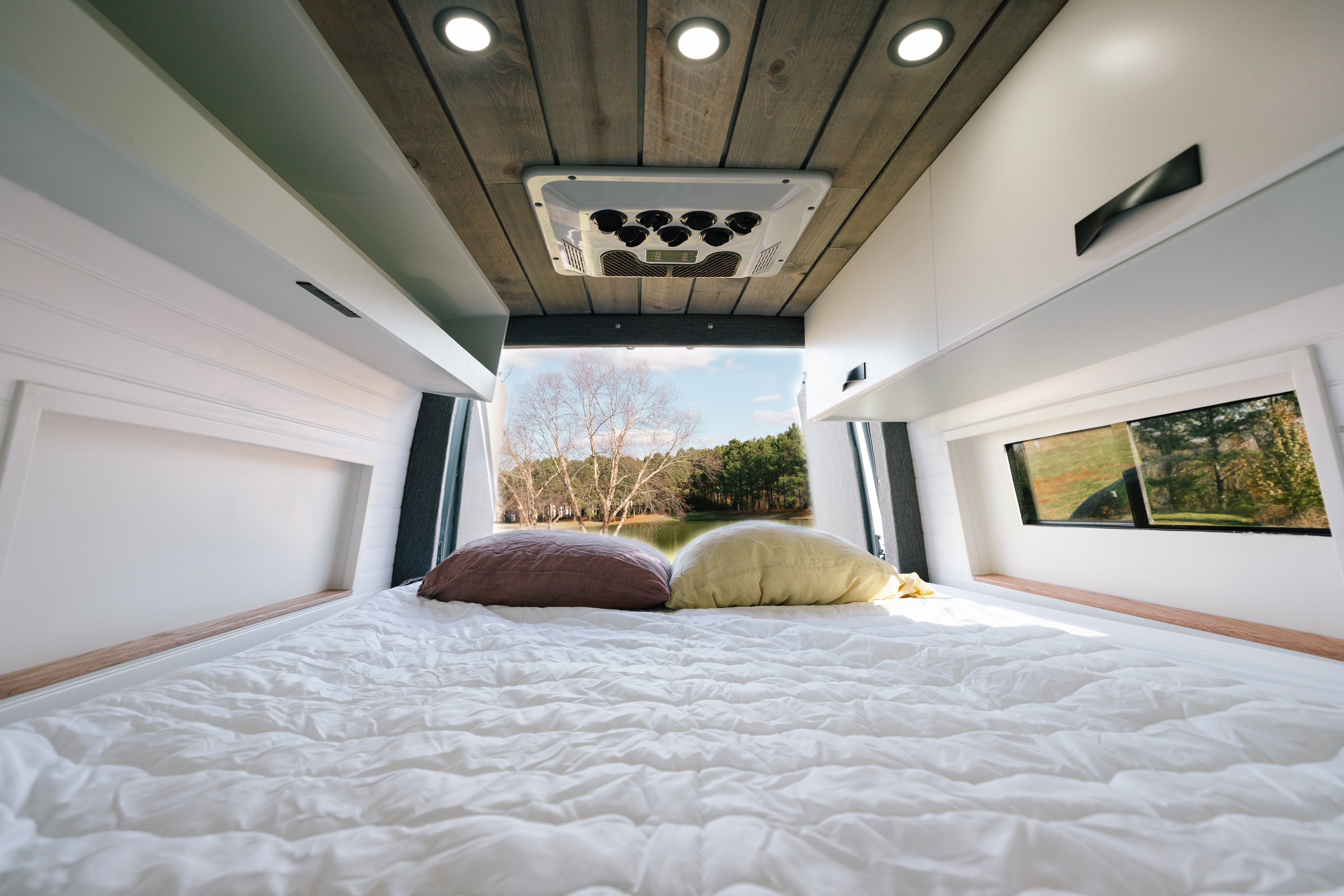 Cozy RV rental interior: white bed, pillows, modern cabinets, and a scenic lake view through the rear window.. Sovereign Vans Mercedes Benz Sprinter 2500 4*4 2024