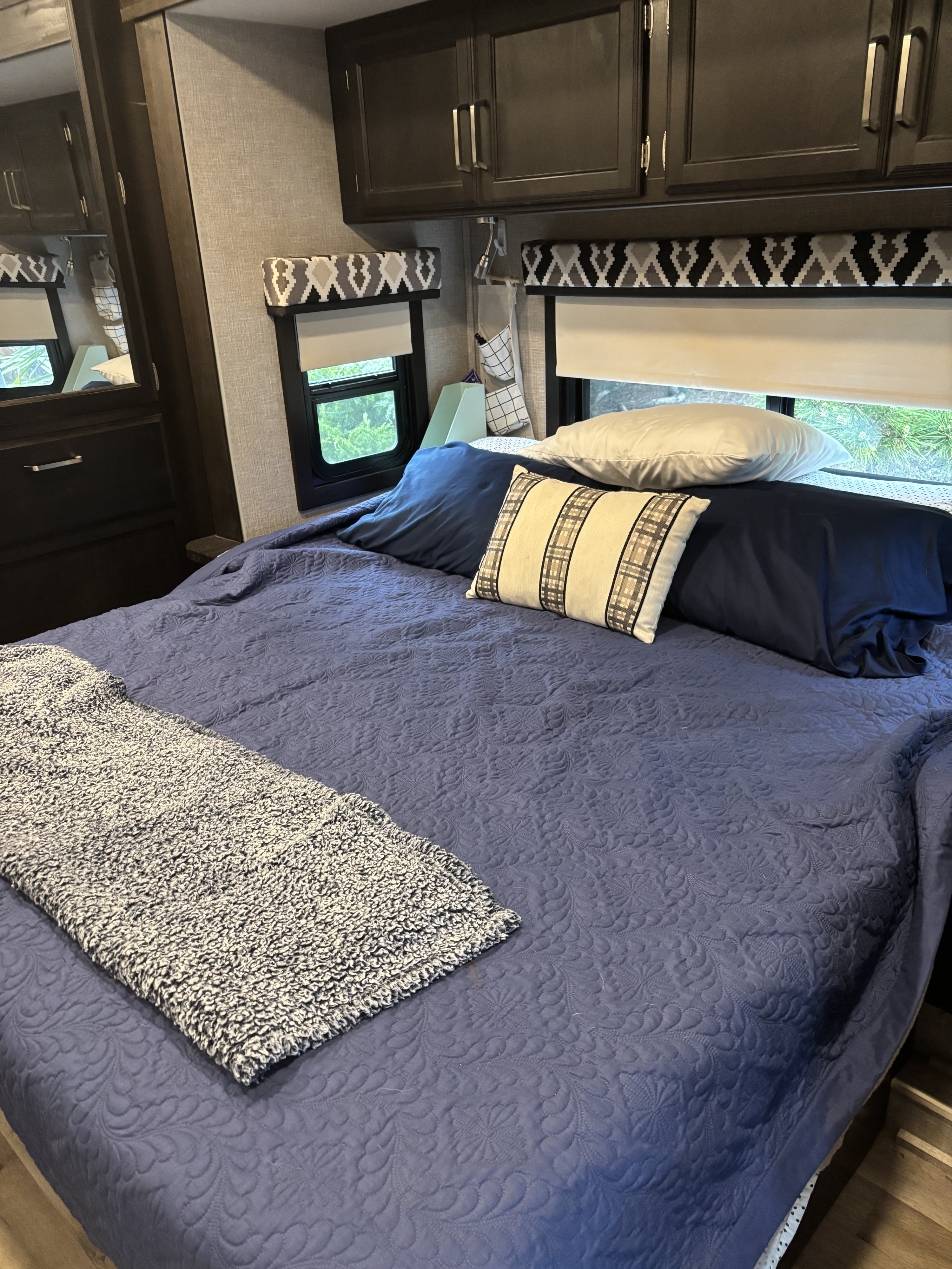 Spacious RV rental bedroom with a blue quilted bed, soft grey throw, dark storage cabinets, and natural light from windows.. Jayco Greyhawk 2022